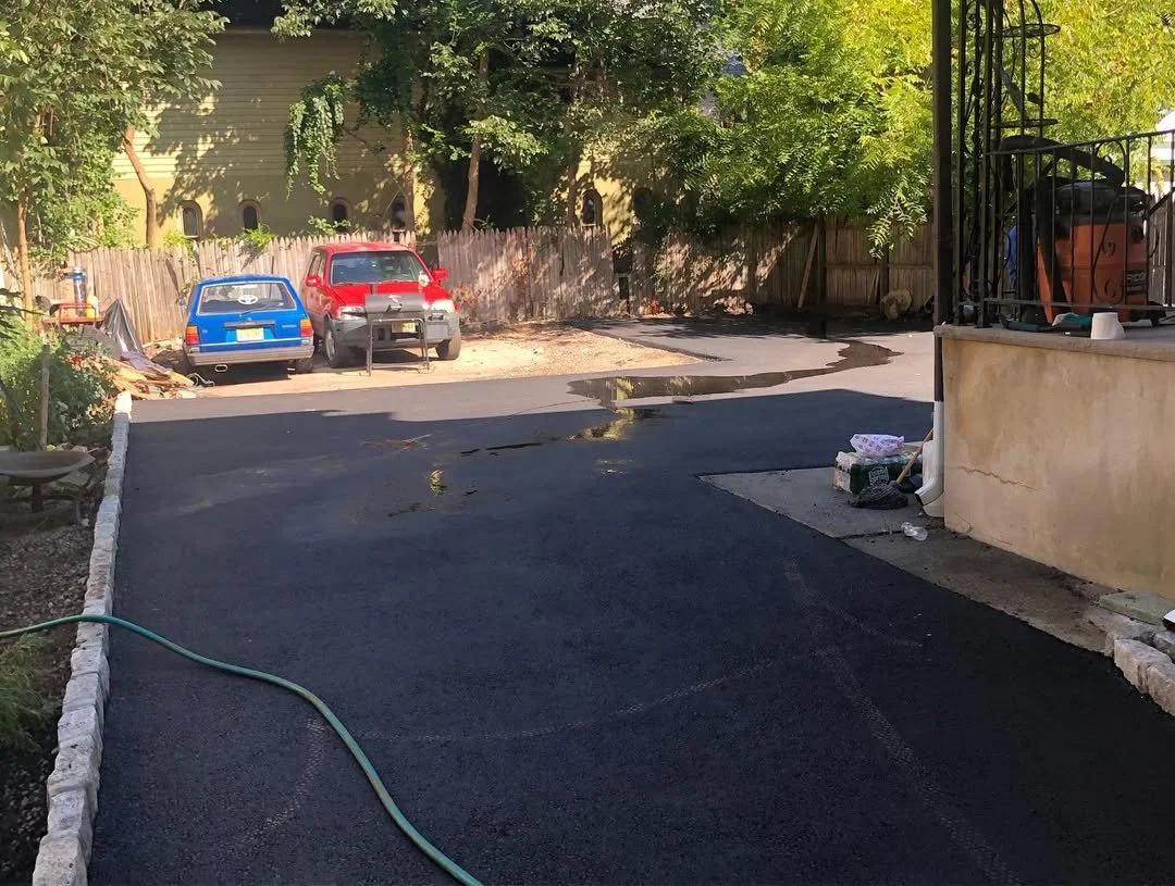 A driveway with fresh asphalt, a garden hose on the ground, and water puddles. Two parked cars, a blue and a red, are visible along with a wooden fence and green leafy trees in the background. There are some objects near the wall and on the porch are