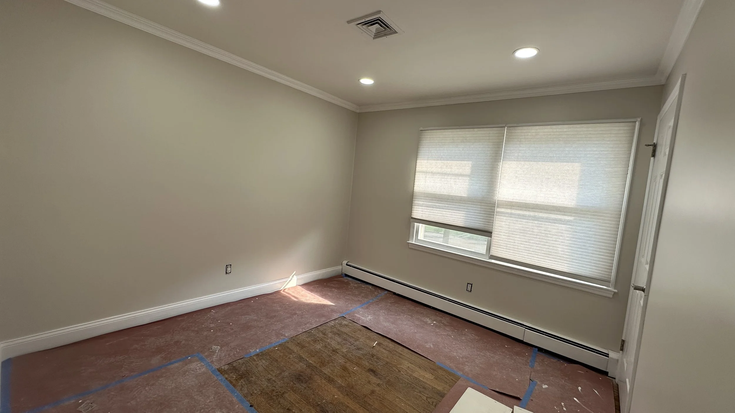 Empty room with beige walls, white ceiling, window with closed blinds, and a partially removed carpet showing hardwood flooring underneath.