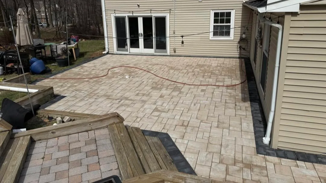 Backyard patio with newly laid light-colored pavers, surrounding a house with beige siding, glass sliding doors, and a small window. Wooden steps lead down to the patio, and there are tools and supplies nearby, indicating ongoing construction or renovation.