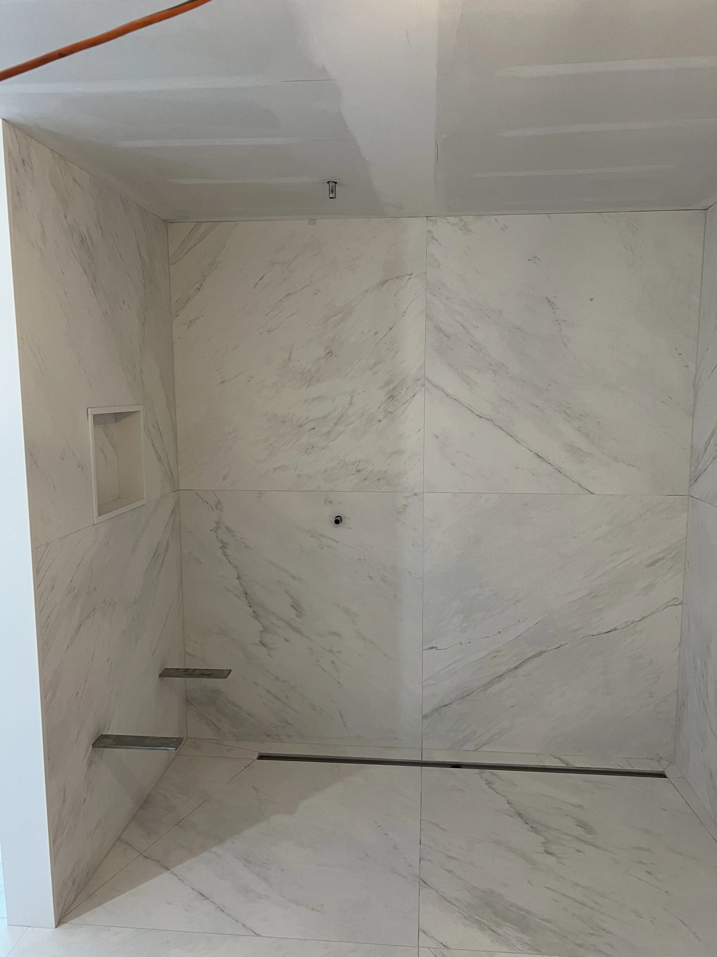 Empty shower with white marble walls, a small built-in shelf, a shower head, a drain, and a glass door.