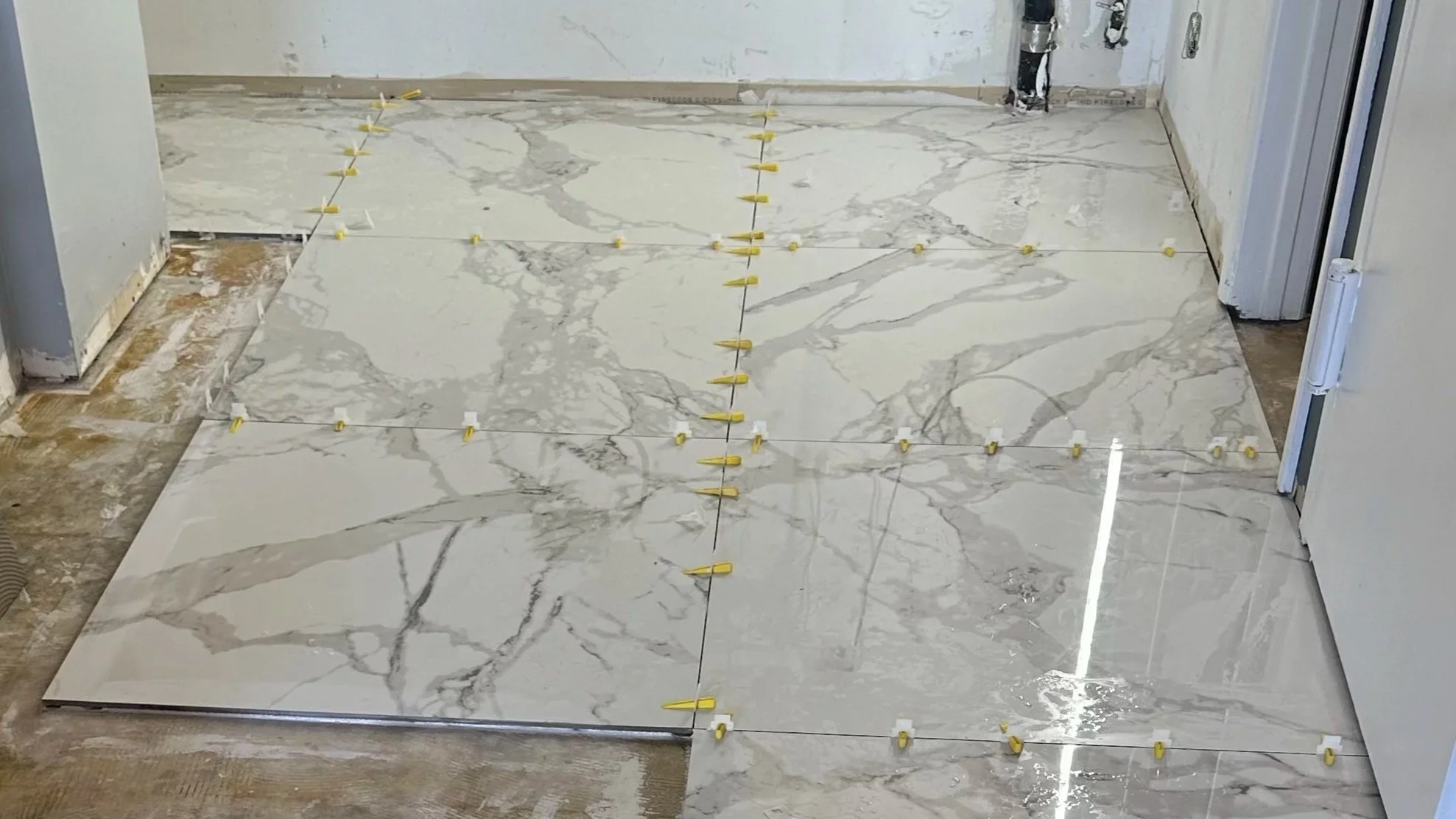 Marble tile flooring being installed in a room, with tiles placed and spaced using yellow spacers, and some parts of the floor not yet tiled.