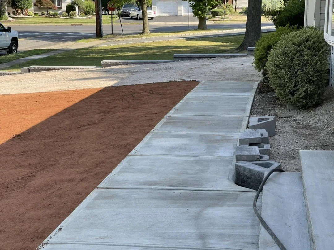 New concrete sidewalk under construction next to a house with bushes, with dirt and gravel areas nearby.
