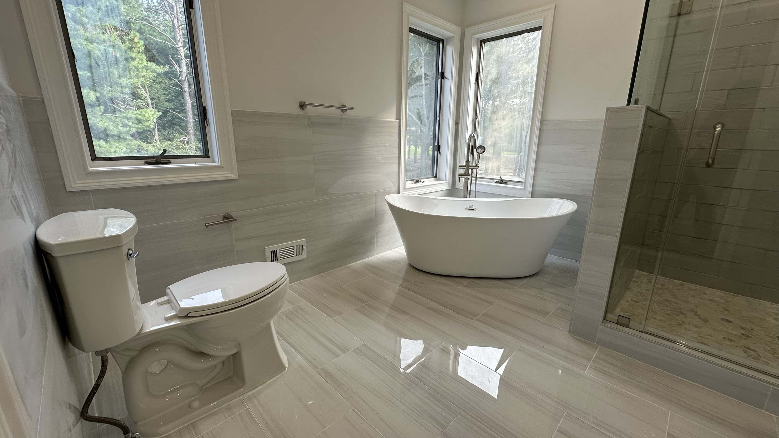 Modern bathroom with a white toilet, a freestanding bathtub, and a glass-enclosed shower. Large windows provide natural light and a view of greenery outside.