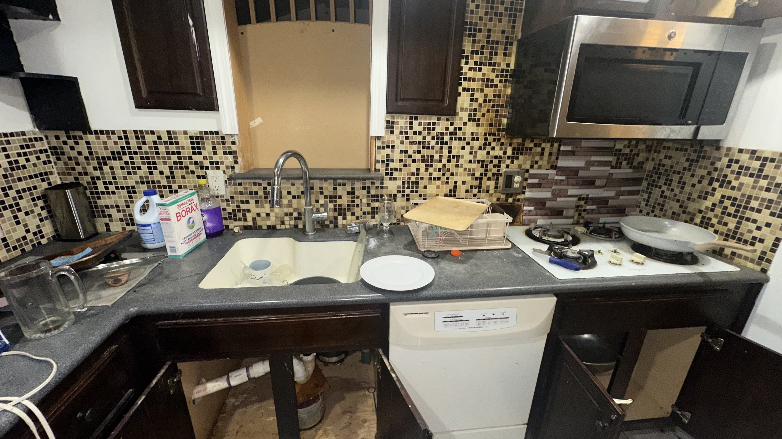 Messy kitchen countertop with a sink, dish soap, cups, plates, and utensils; stove with a frying pan; microwave; cabinets; backsplash with small mosaic tiles; open cabinet drawers with cleaning supplies underneath.