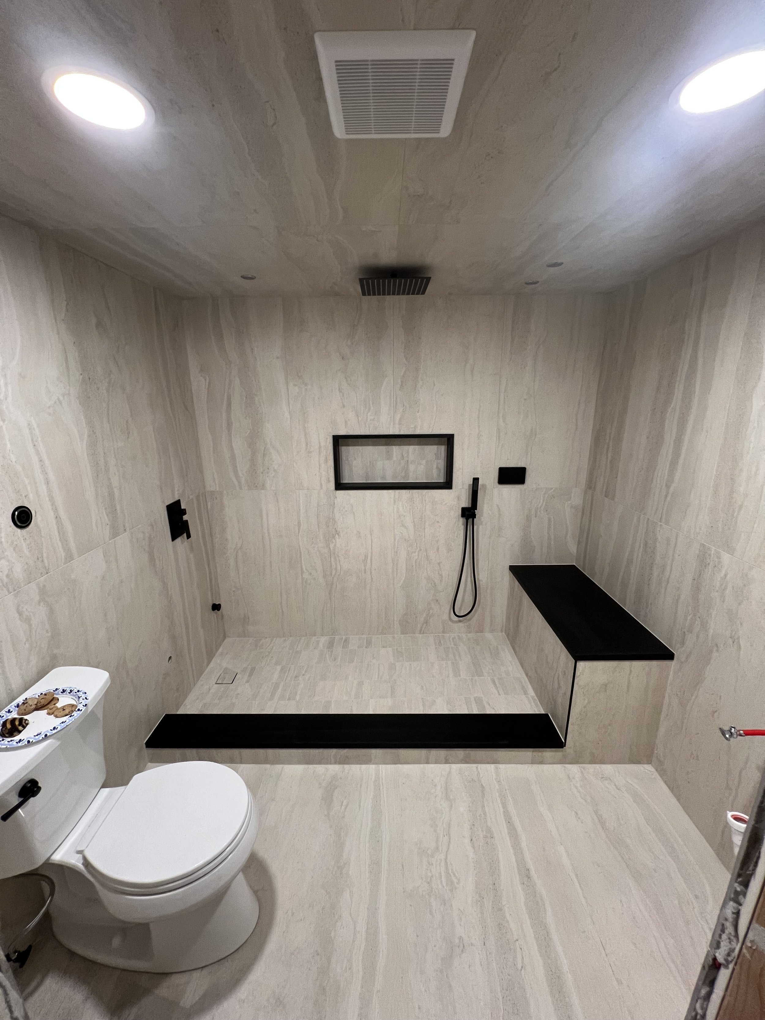 Modern bathroom with beige tiles, a toilet, a black shelf, a black mirror frame, a black shower head, and a black vent on the ceiling. Small window near the ceiling.