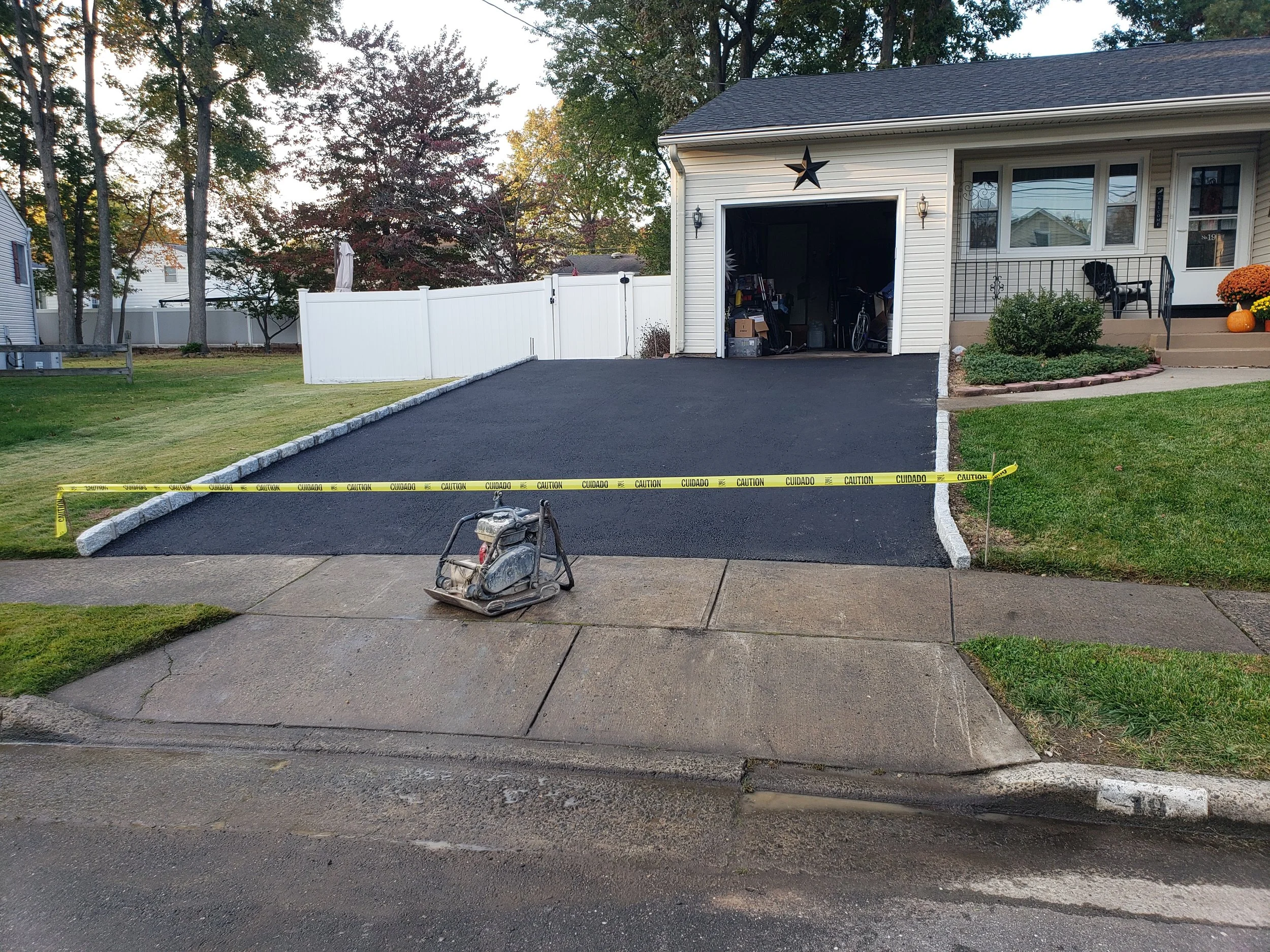 Newly paved black asphalt driveway in front of a house with a garage door, caution tape blocking access, a sidewalk, and a weed trimmer on the driveway.