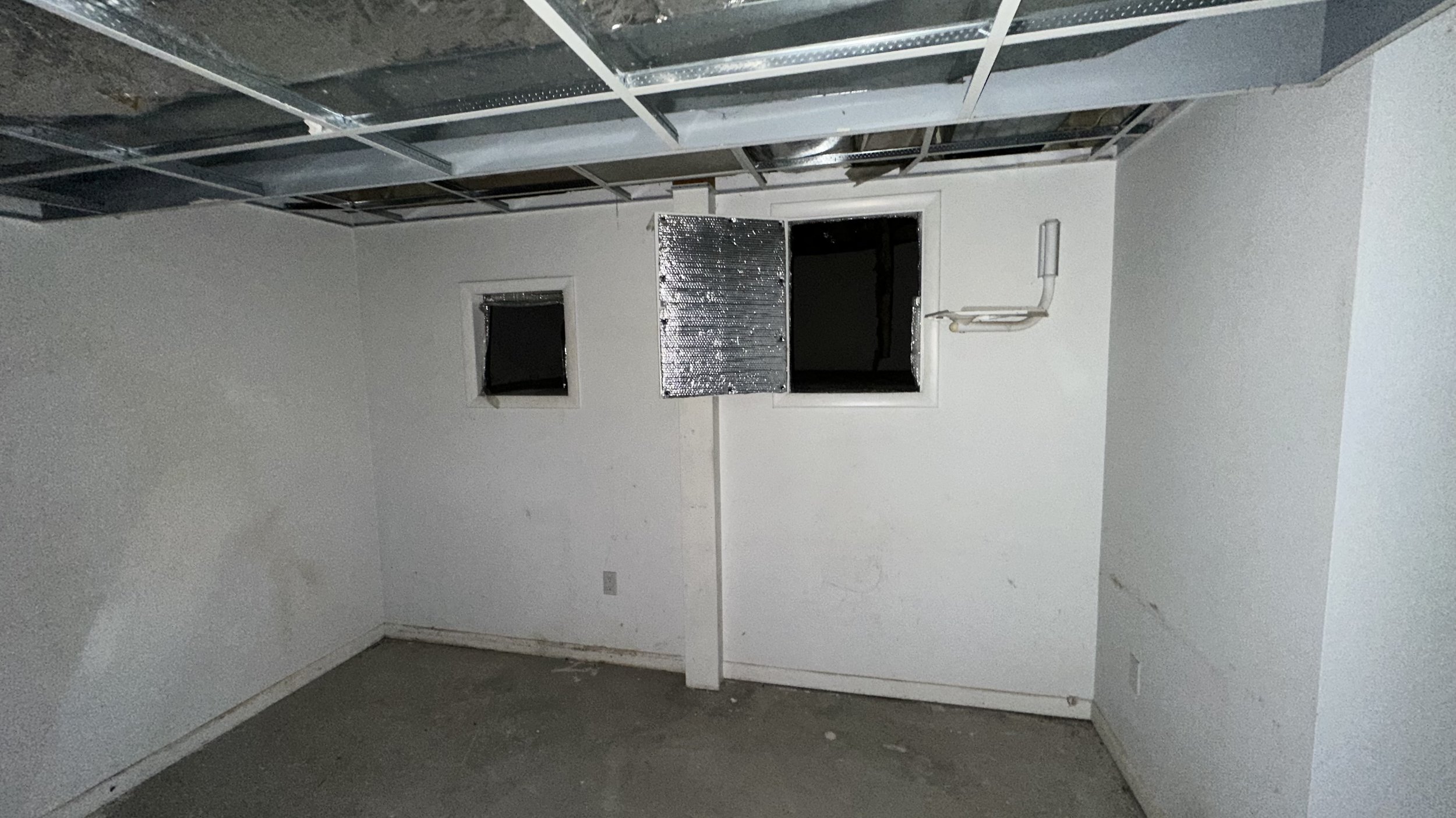 A small unfinished room with white walls, an open window with a metal shutter, and exposed ceiling rafters and ductwork.