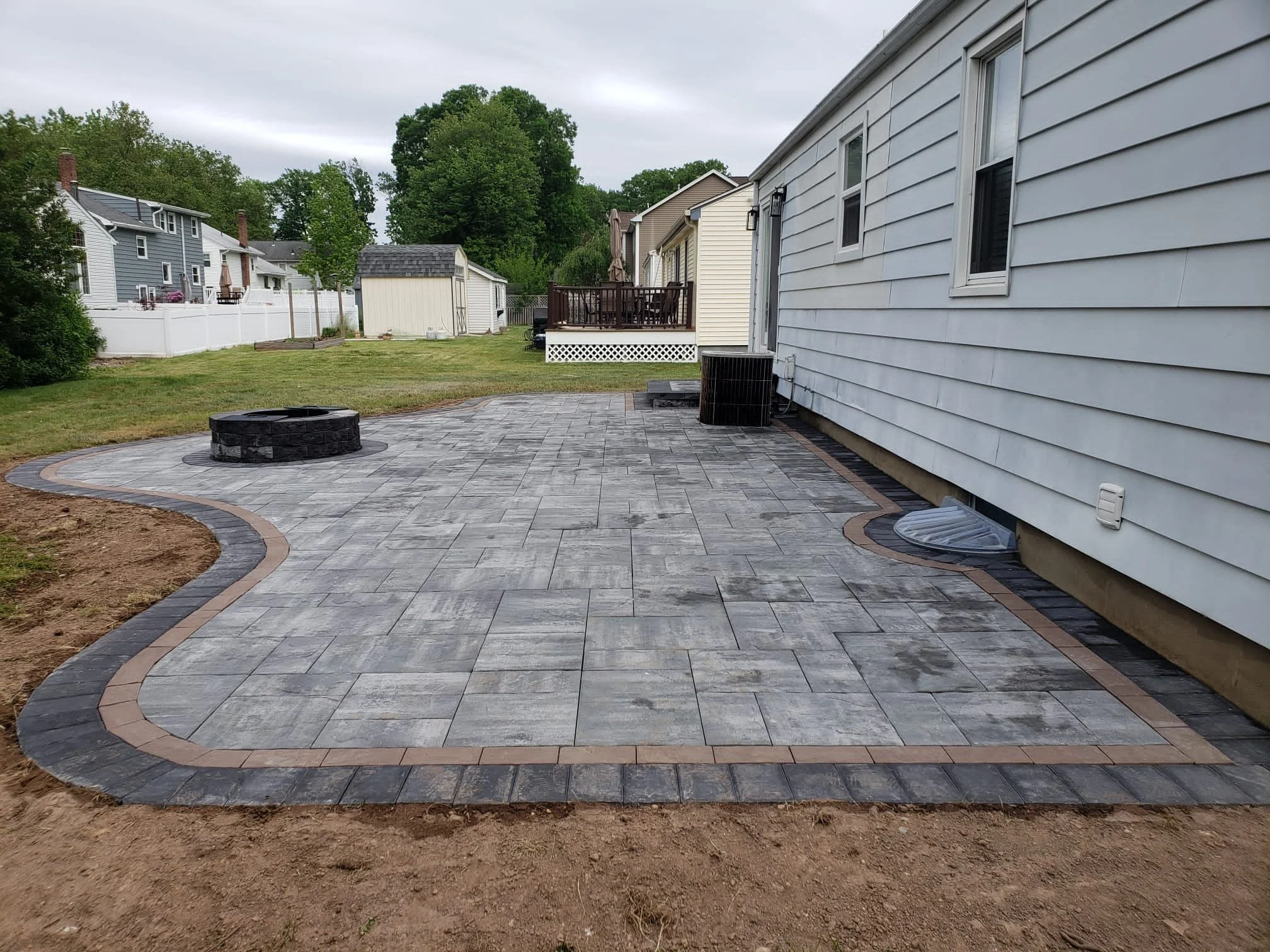 Newly paved backyard patio with gray stones and a circular fire pit in a suburban backyard.