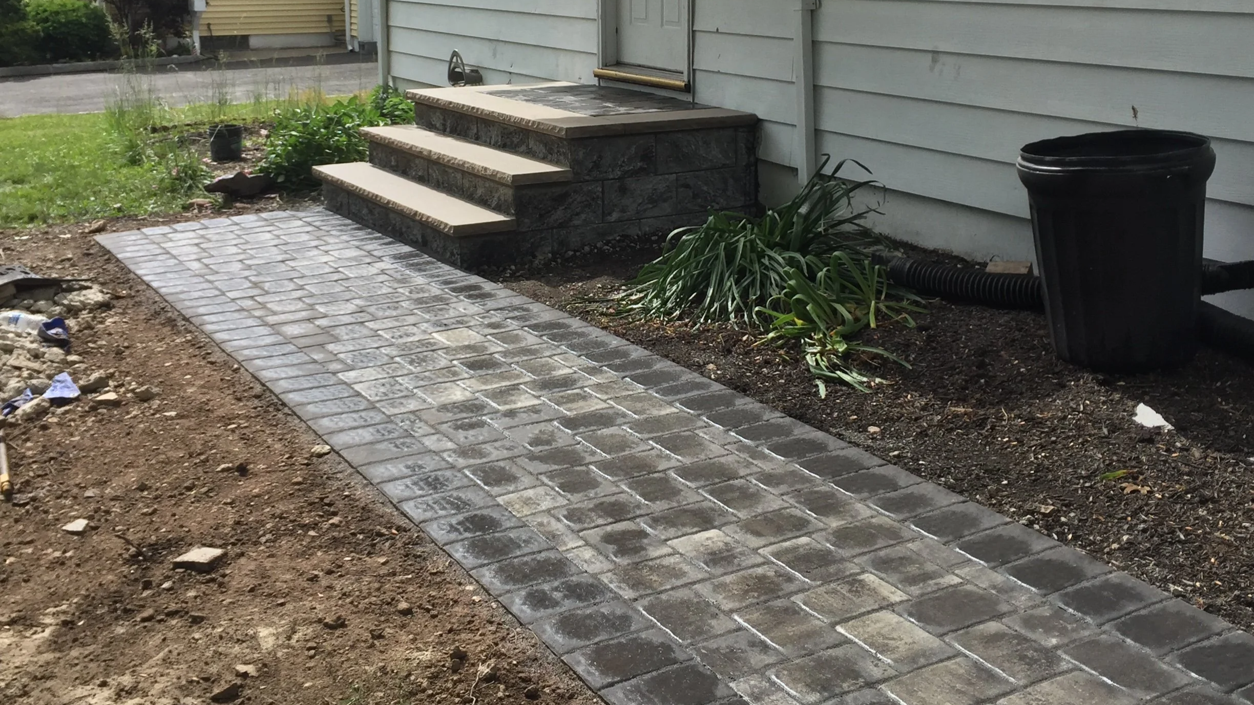 Newly paved stone walkway leading to the front steps of a house, with a small garden bed to the side and a black trash can near the house wall.