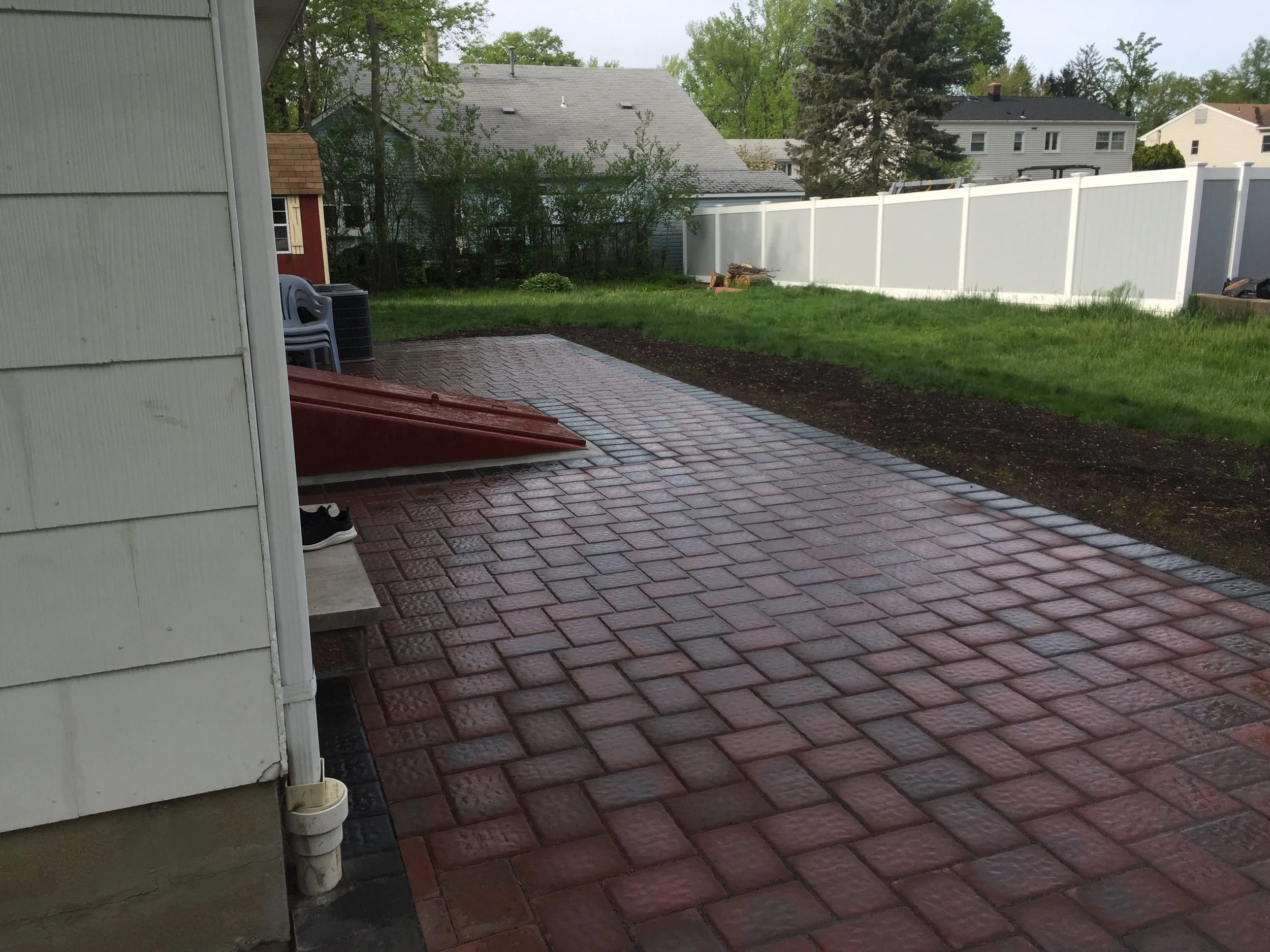 Backyard with freshly laid reddish-brown brick patio, white fenced yard, green grass, and neighboring houses in the background.