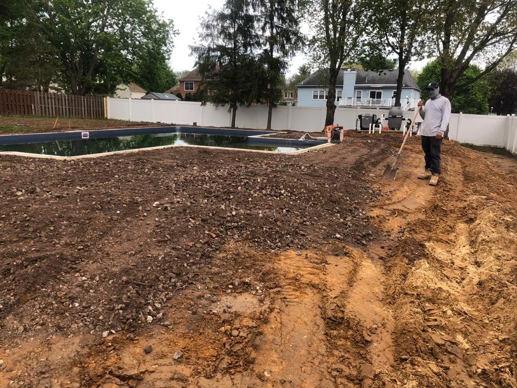 A man with a face mask and a hat shoveling dirt in a backyard near a pool that is under construction, with construction equipment and white fences in the background.