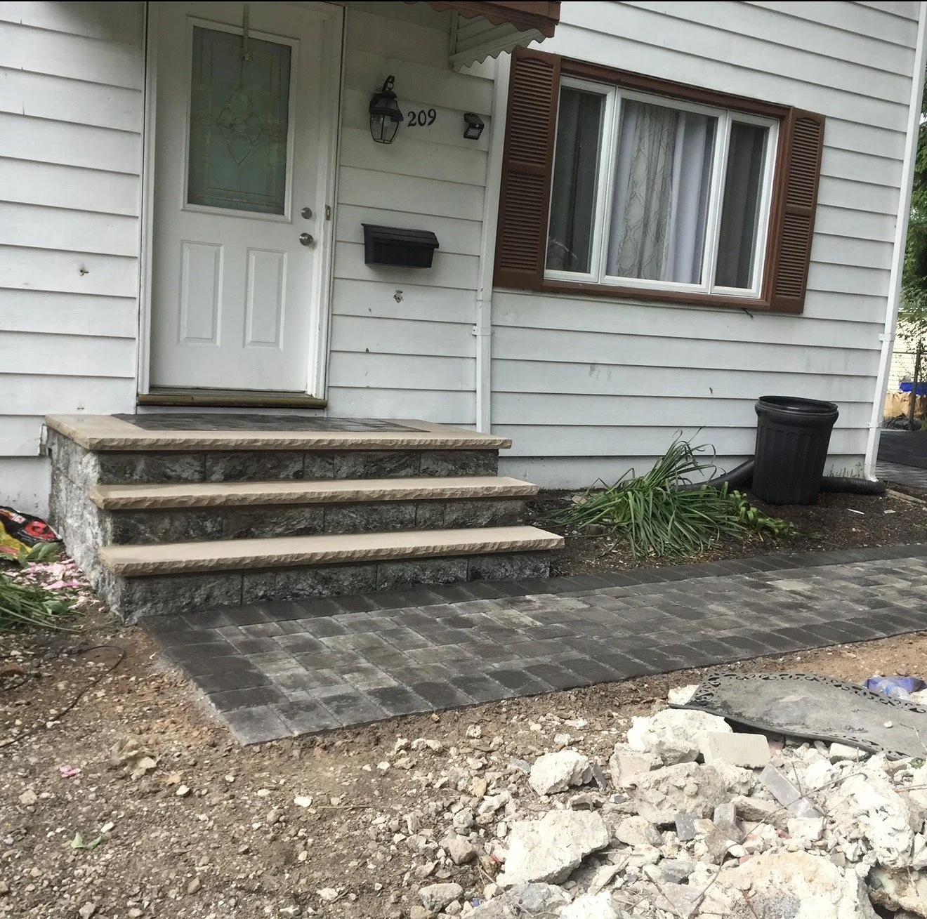 Front entrance of a house with three steps leading up to the white door, window with brown shutters, a black mailbox, black trash can, and landscaping with plants and paving stones, with construction debris in the foreground.