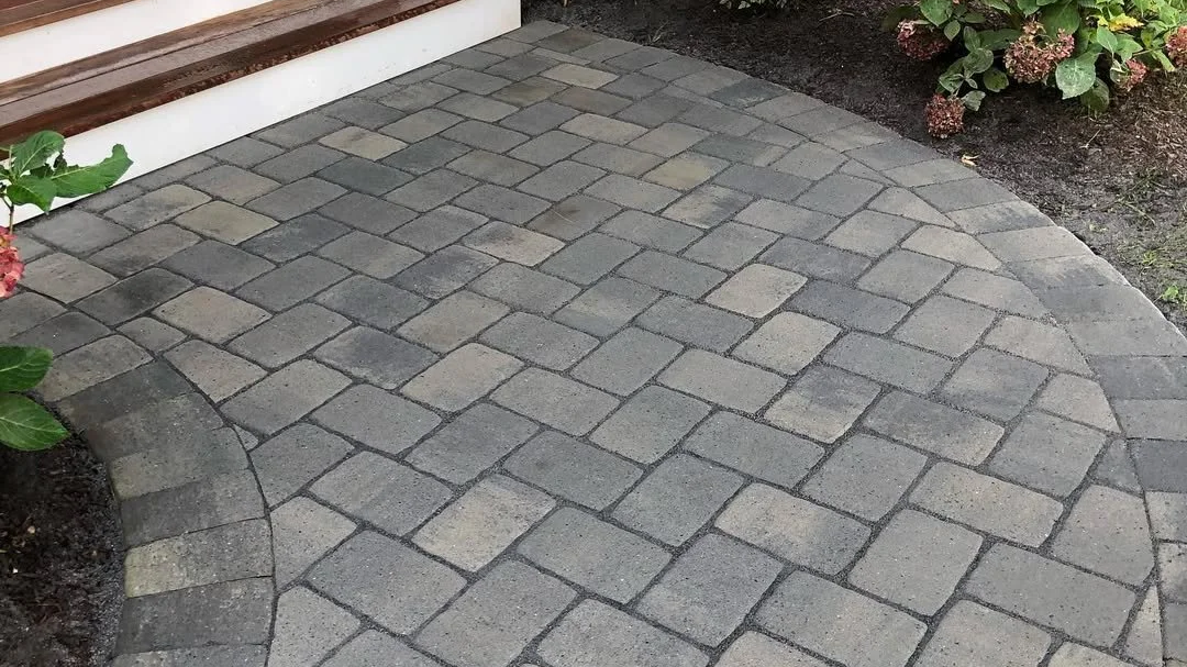 Curved stone paver walkway with gray and beige bricks near a garden bed with pink flowers and green leaves.