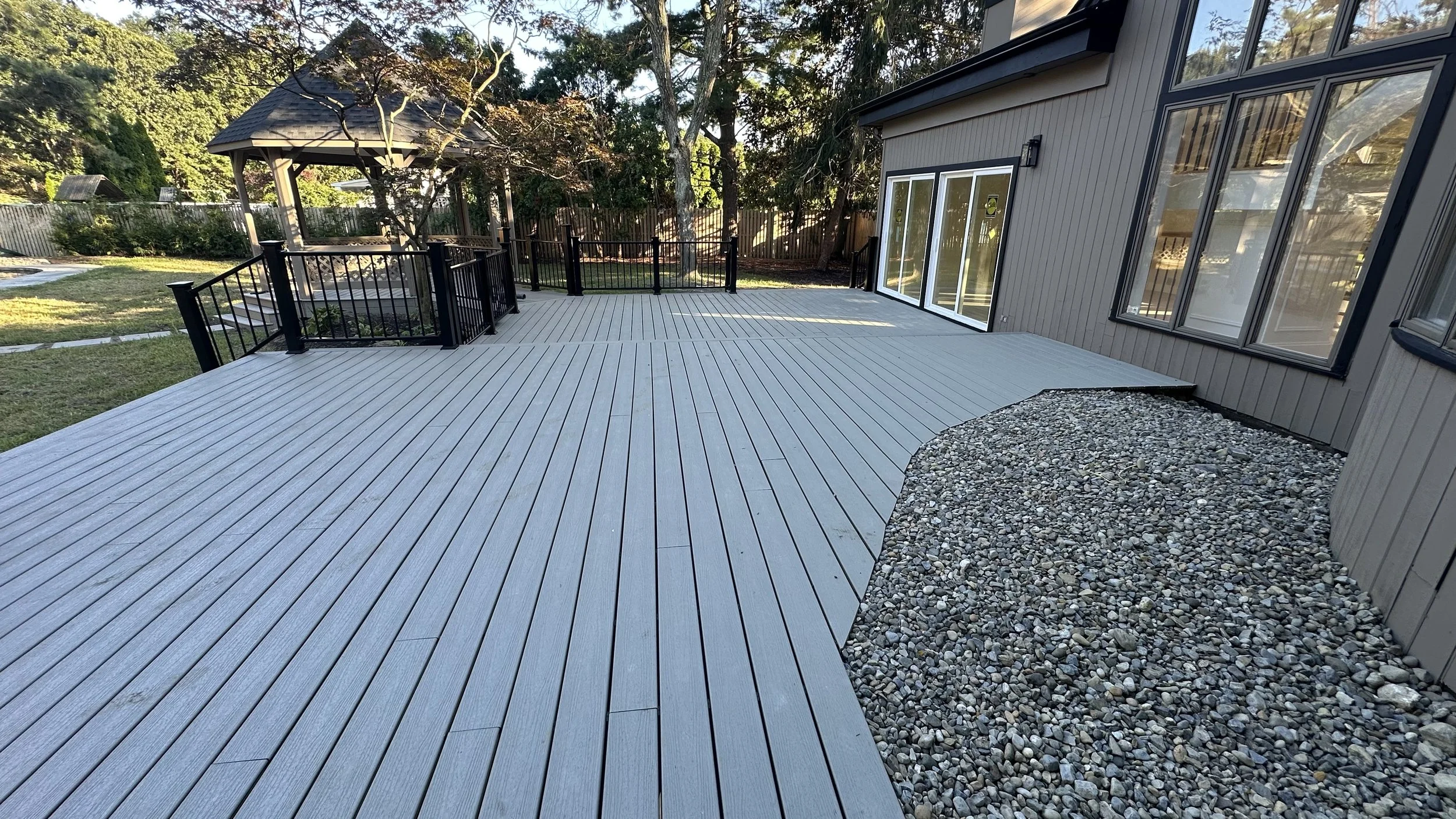 Spacious outdoor deck with light-colored wood planking, surrounded by a black metal fence, next to a house with gray siding and large windows. A small gravel area borders the deck, and there is a gazebo with a shingled roof visible in the background among trees and green lawn.