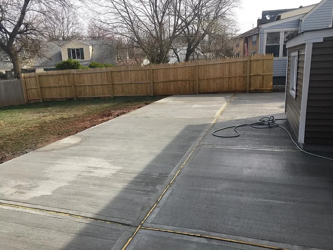 A newly poured concrete backyard patio with a wooden fence in the background and a house on the right side, with a garden hose coiled on the side of the house.