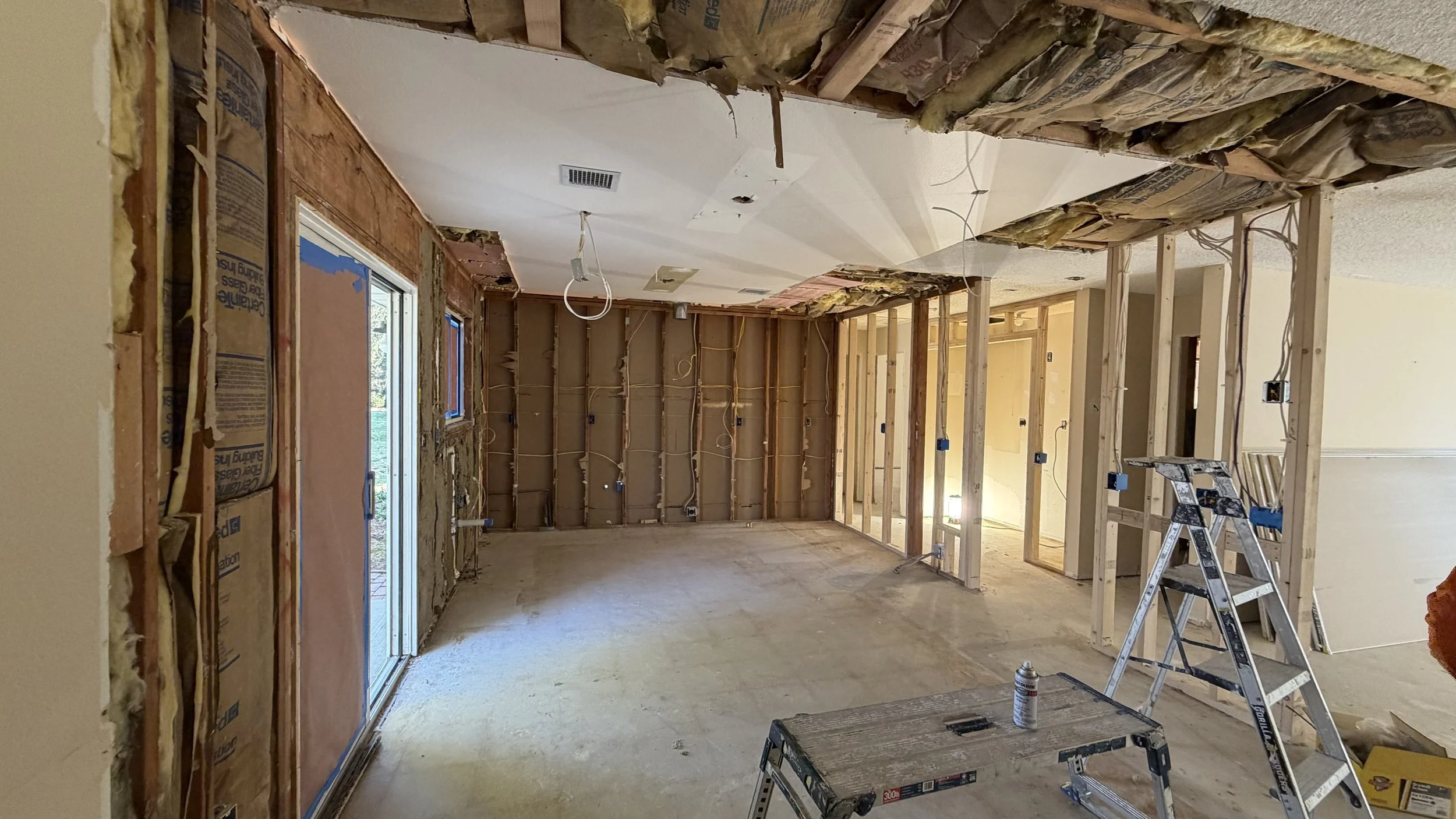 Interior of a house under renovation with exposed wooden framing, cut-out sections, and construction tools. A sliding glass door is visible on the left side, leading outside. The ceiling shows insulation and missing drywall sections. Walls are partially open, revealing internal wiring.