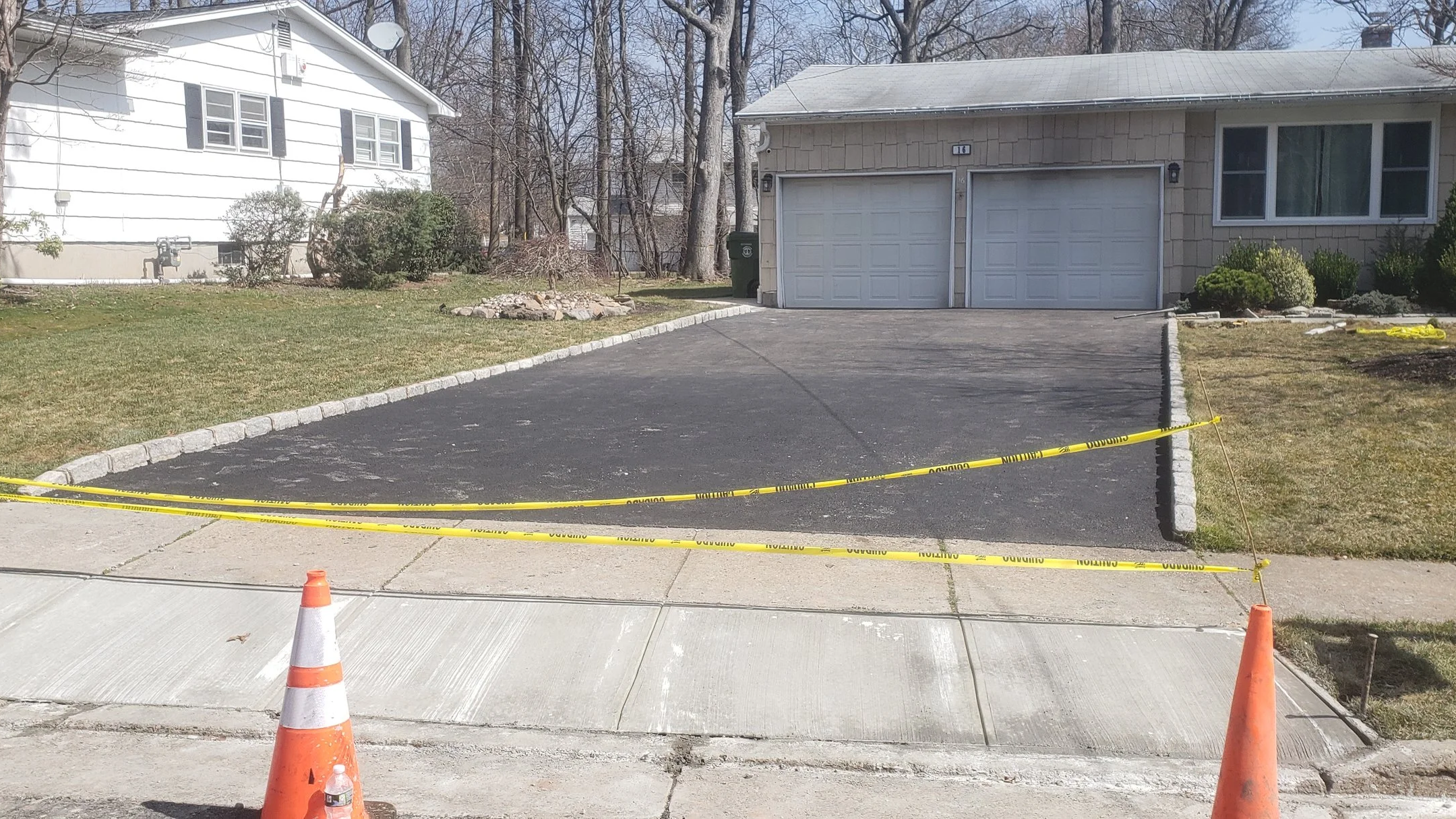 A driveway section is blocked off with yellow caution tape and orange cones, in front of a house with a two-car garage and a front yard with grass and bushes.