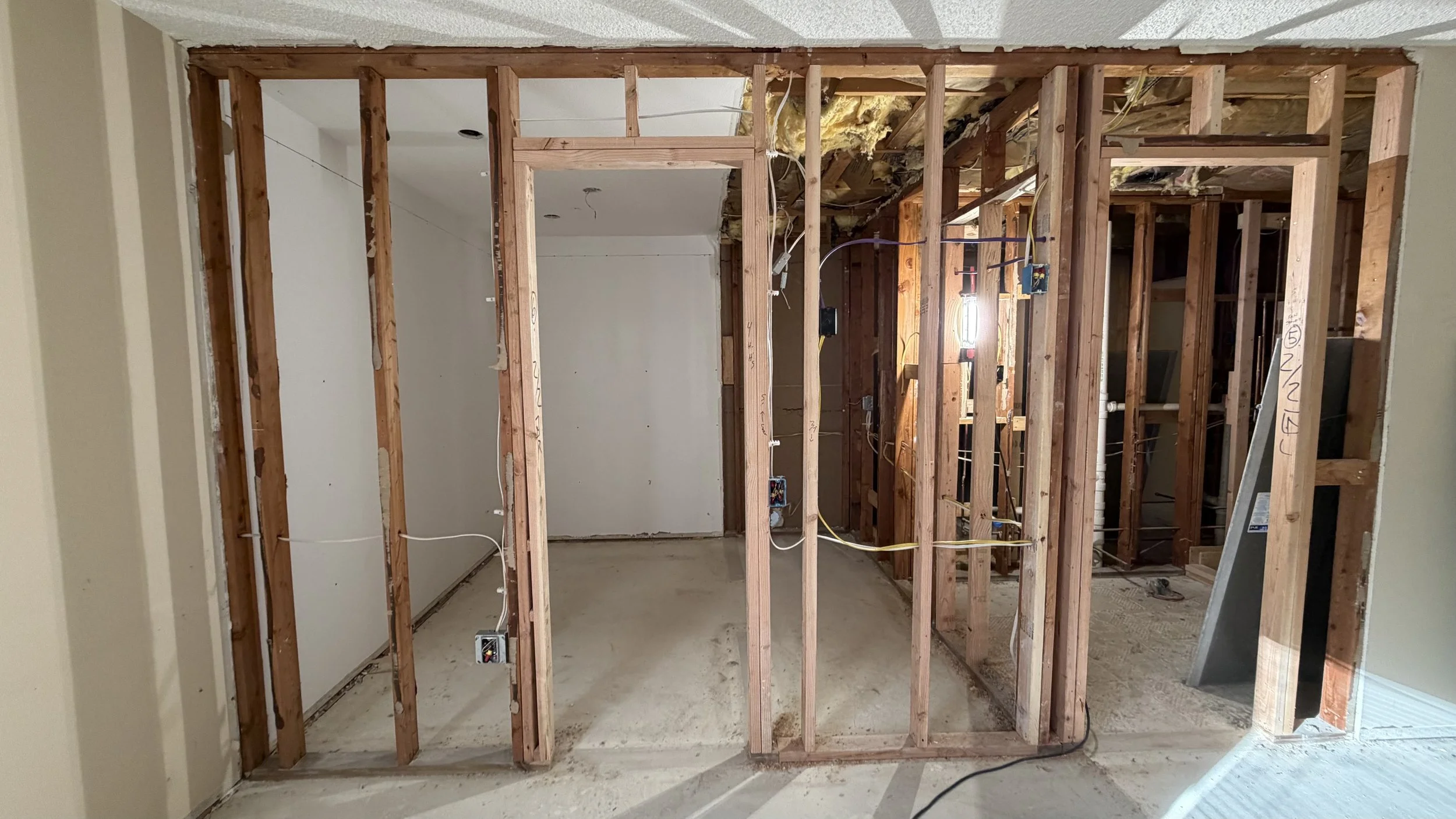 Interior view of a home under renovation showing wooden framing, electrical wiring, and exposed insulation, with a partially finished floor and an open doorway leading to another room.