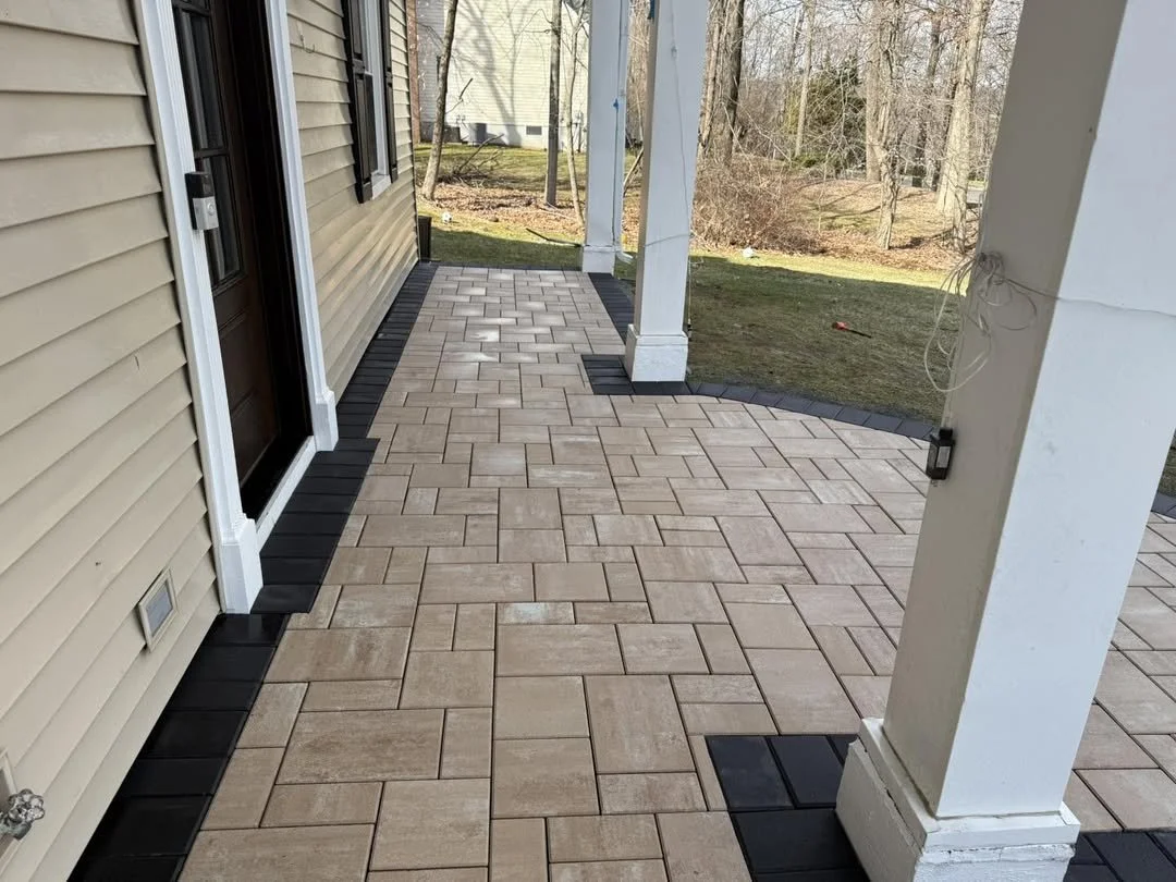 A backyard patio with beige brick pavers, black border tiles, and a house with beige siding, black shutters, and a screened door. There is a grassy yard and trees in the background.