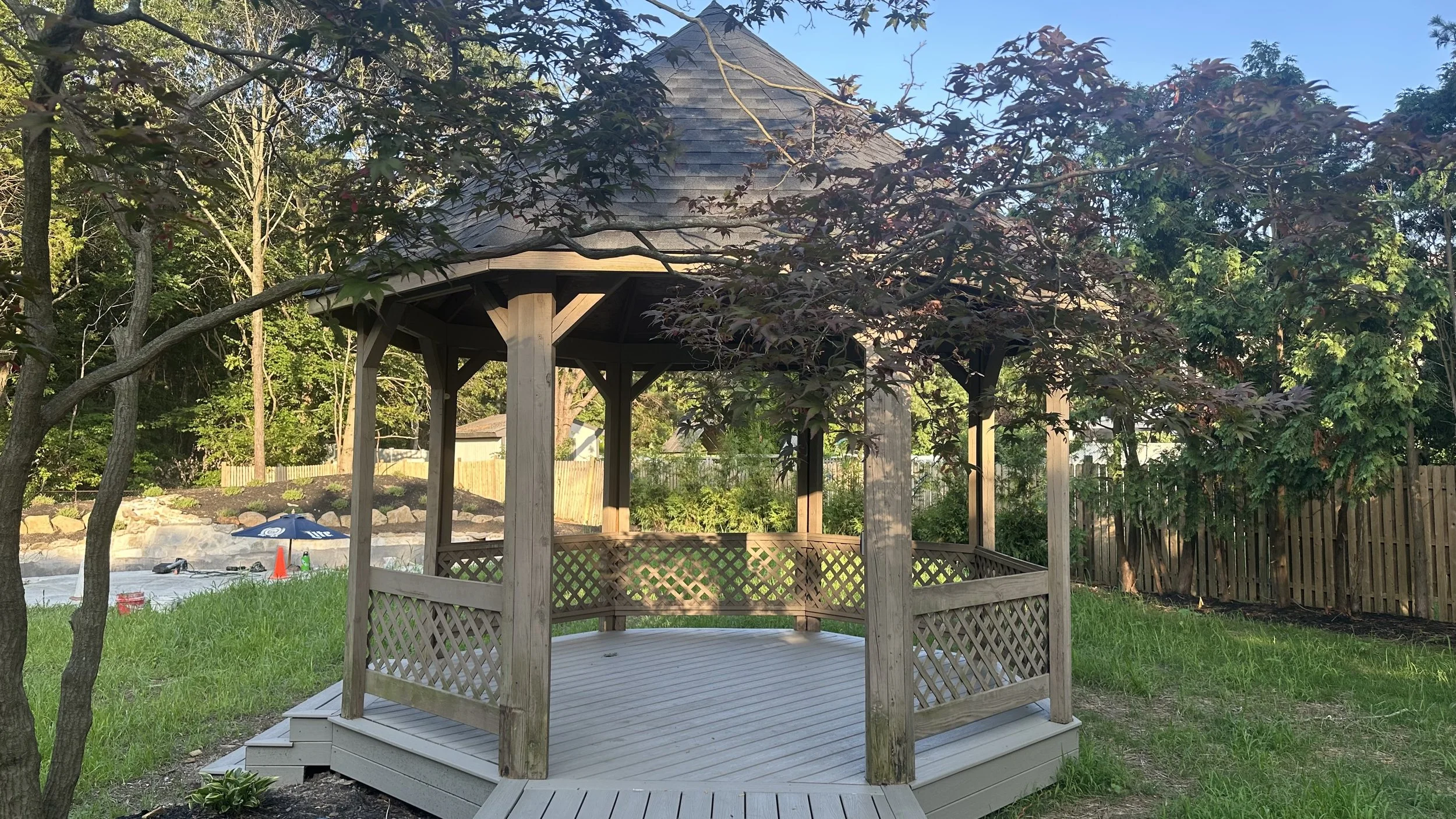 Wooden gazebo in a grassy backyard with trees and a wooden fence in the background.