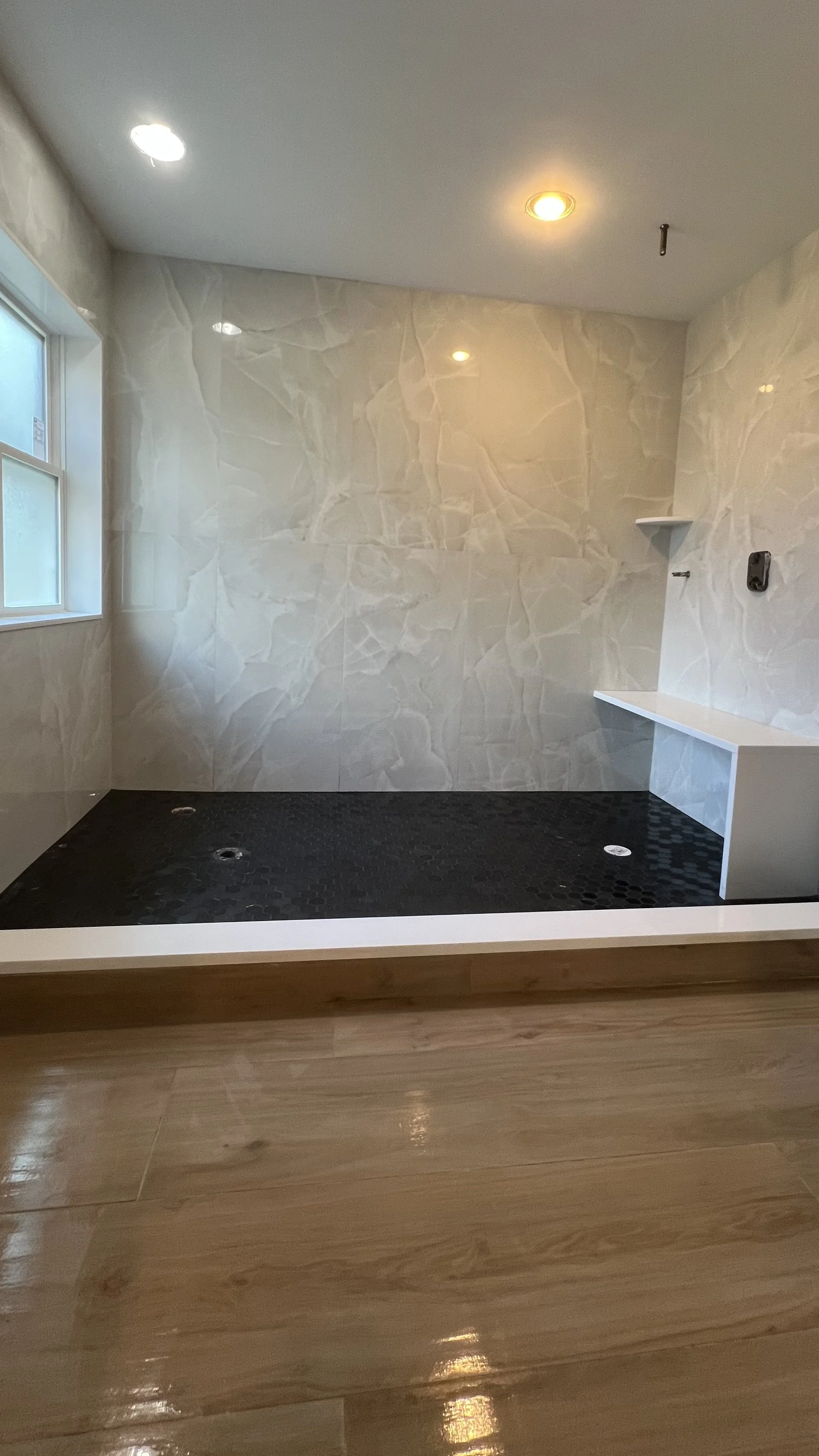 Empty bathroom shower area with marble walls, a window, two recessed ceiling lights, and a black hex tile floor with drain holes.