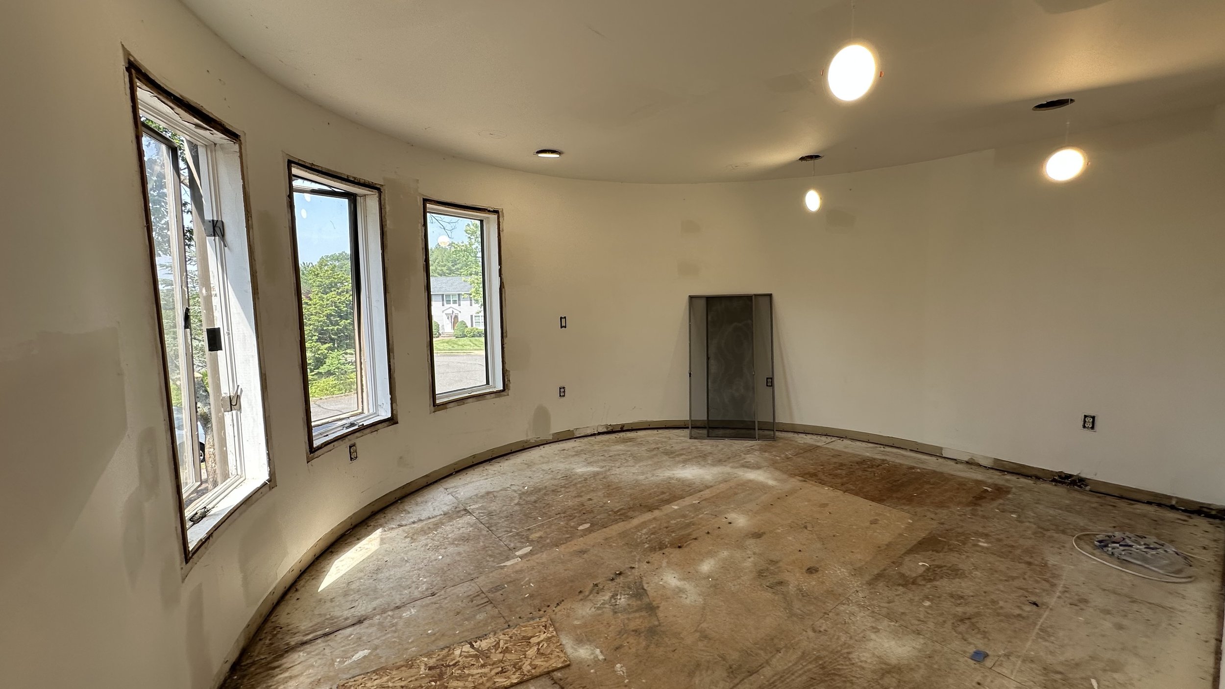 Interior of a room under renovation with three windows, unfinished floor, and ceiling lights.