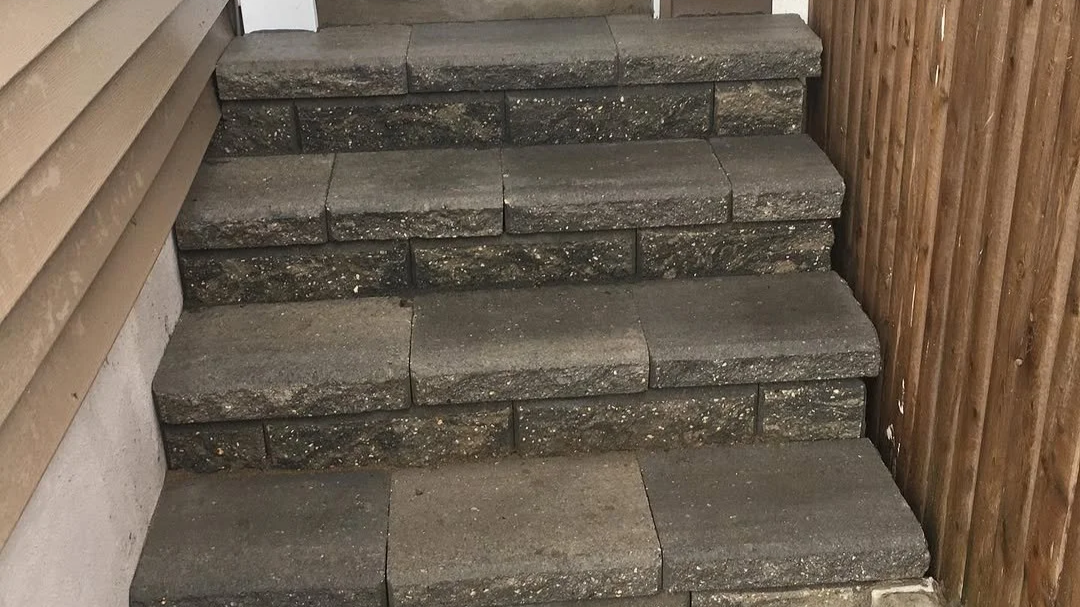 Three concrete steps with a black and gray pattern, flanked by a wooden fence on the right and a wall on the left.