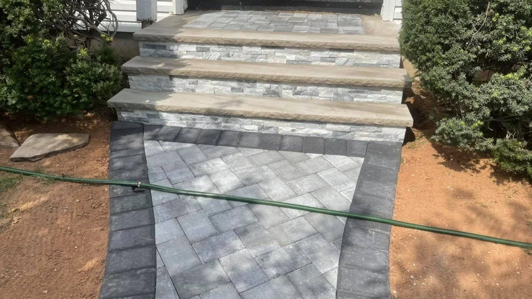 Stone steps leading to a porch with a patterned stone walkway in front, surrounded by landscaped area with bushes and soil.