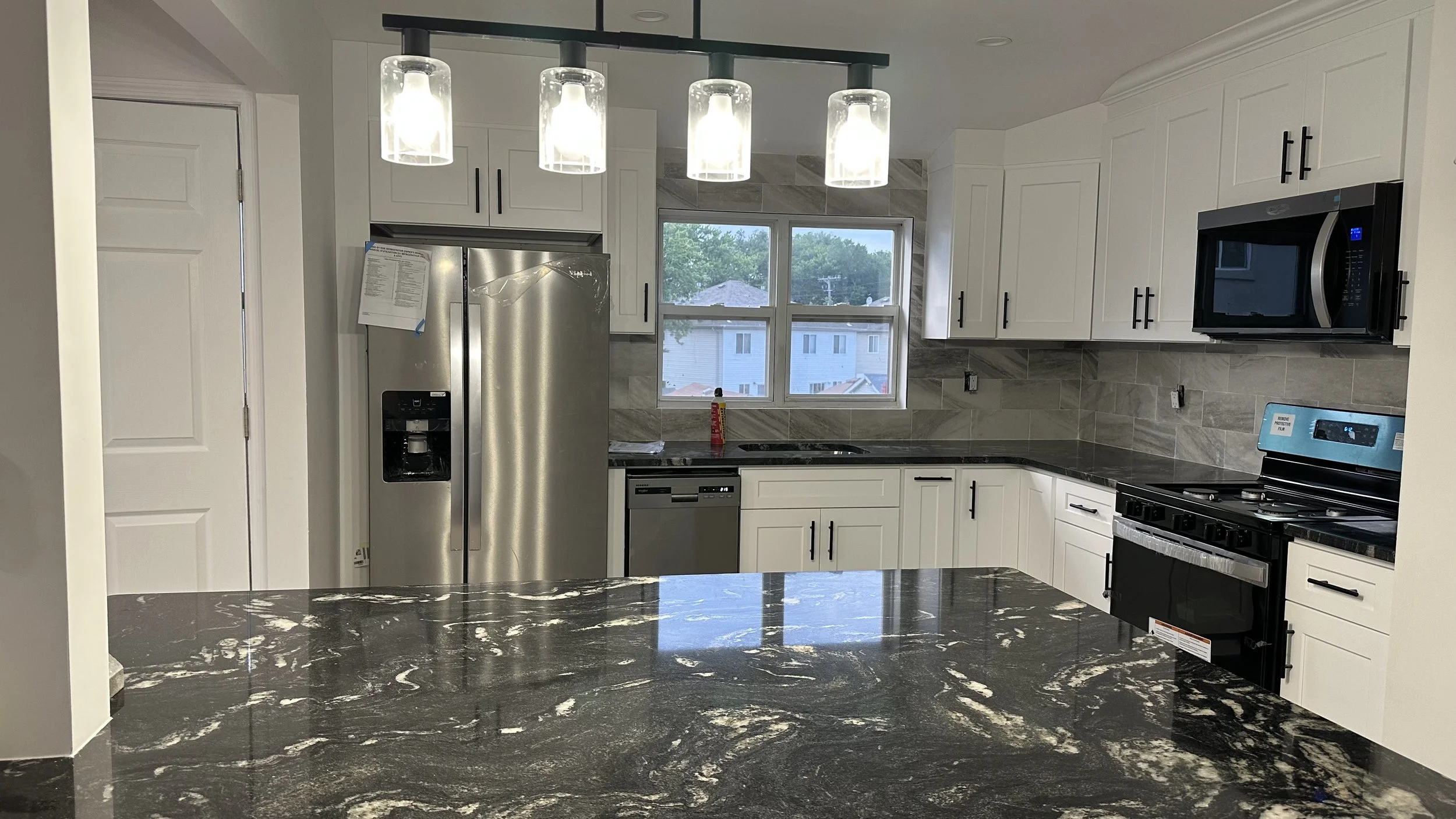 Modern kitchen with white cabinets, black hardware, granite countertops, stainless steel refrigerator, microwave, dishwasher, and stove, with a window showing neighboring houses outside, and four pendant lights hanging above the island.
