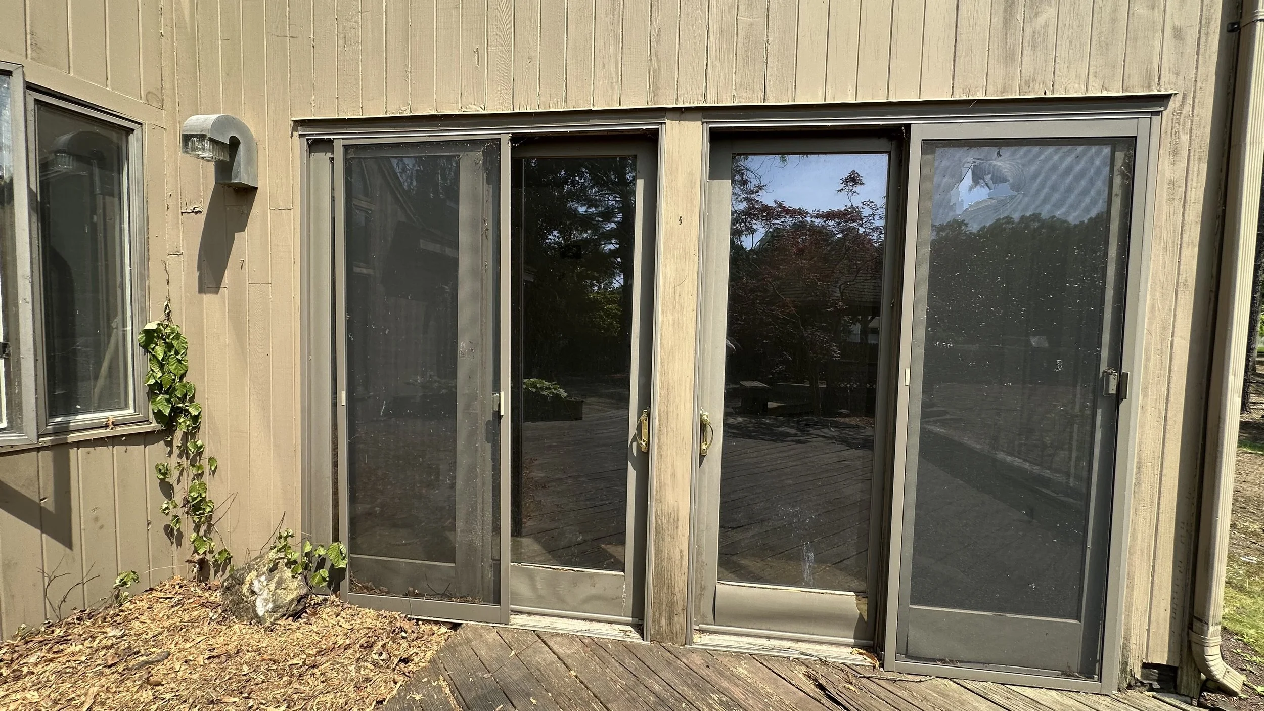 Exterior view of a building with sliding glass doors, a window with a screen, an outdoor light fixture, and some ivy climbing the wall, on a wooden deck.