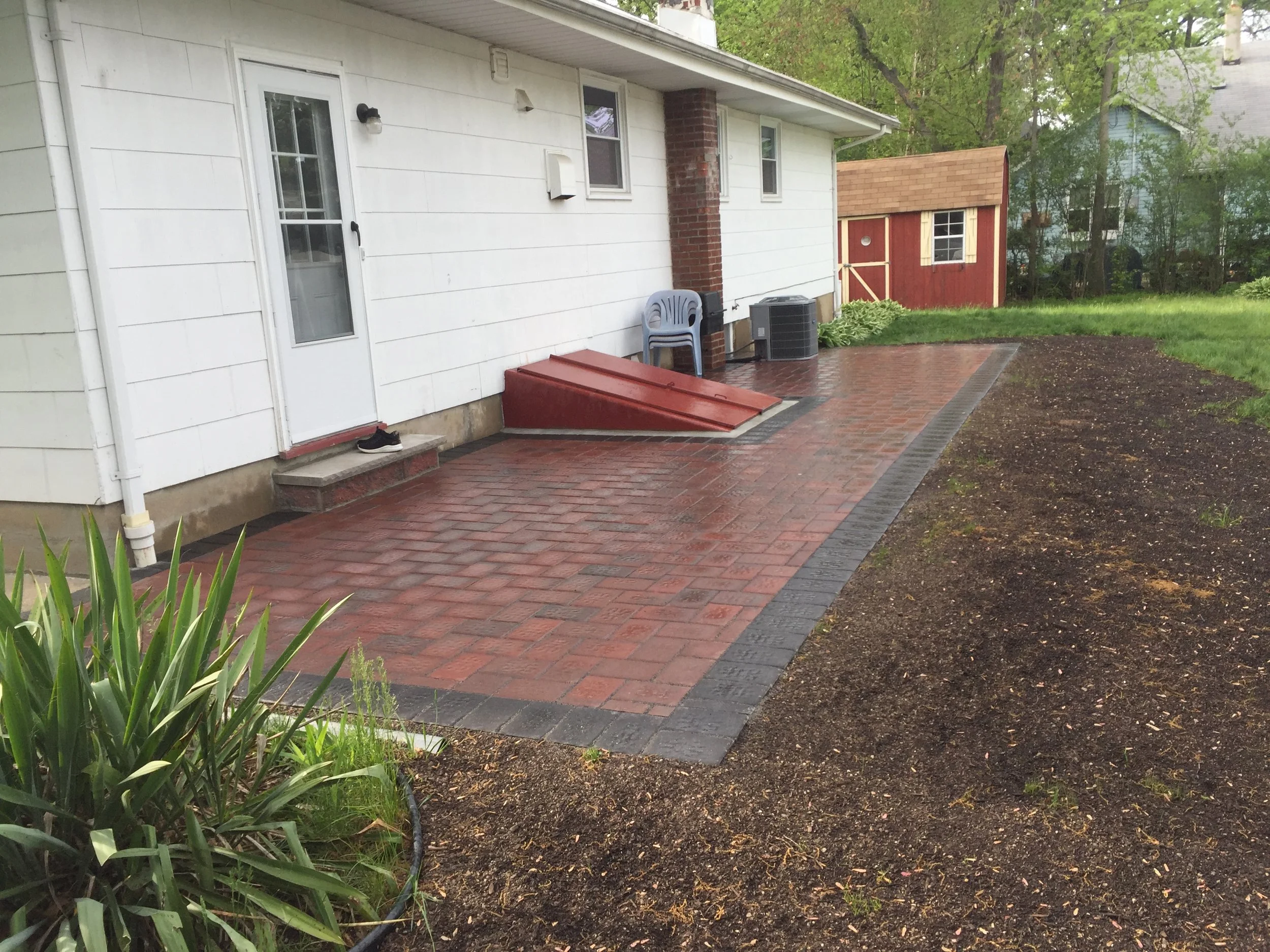 Backyard patio with red and black brick pavers, a white house with a door and window, a small shed, and some garden plants.