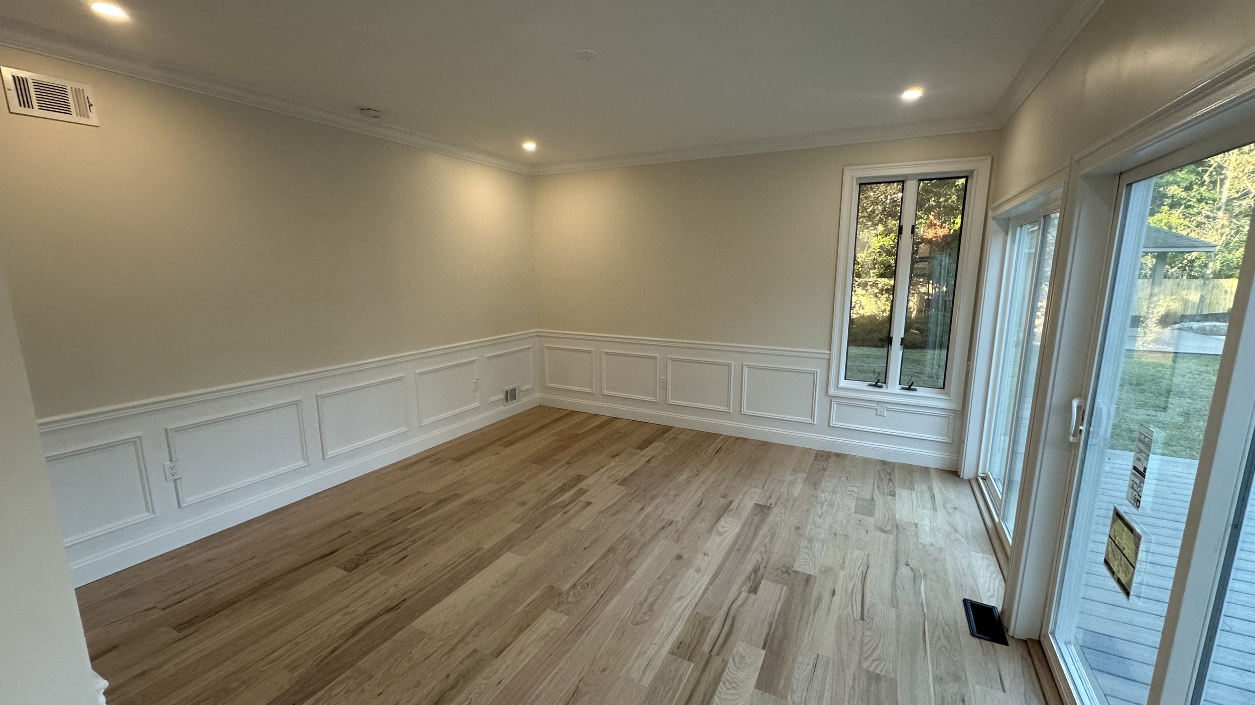 Empty room with hardwood flooring, white walls, decorative wainscoting, natural light from large windows and sliding glass door to the outside.