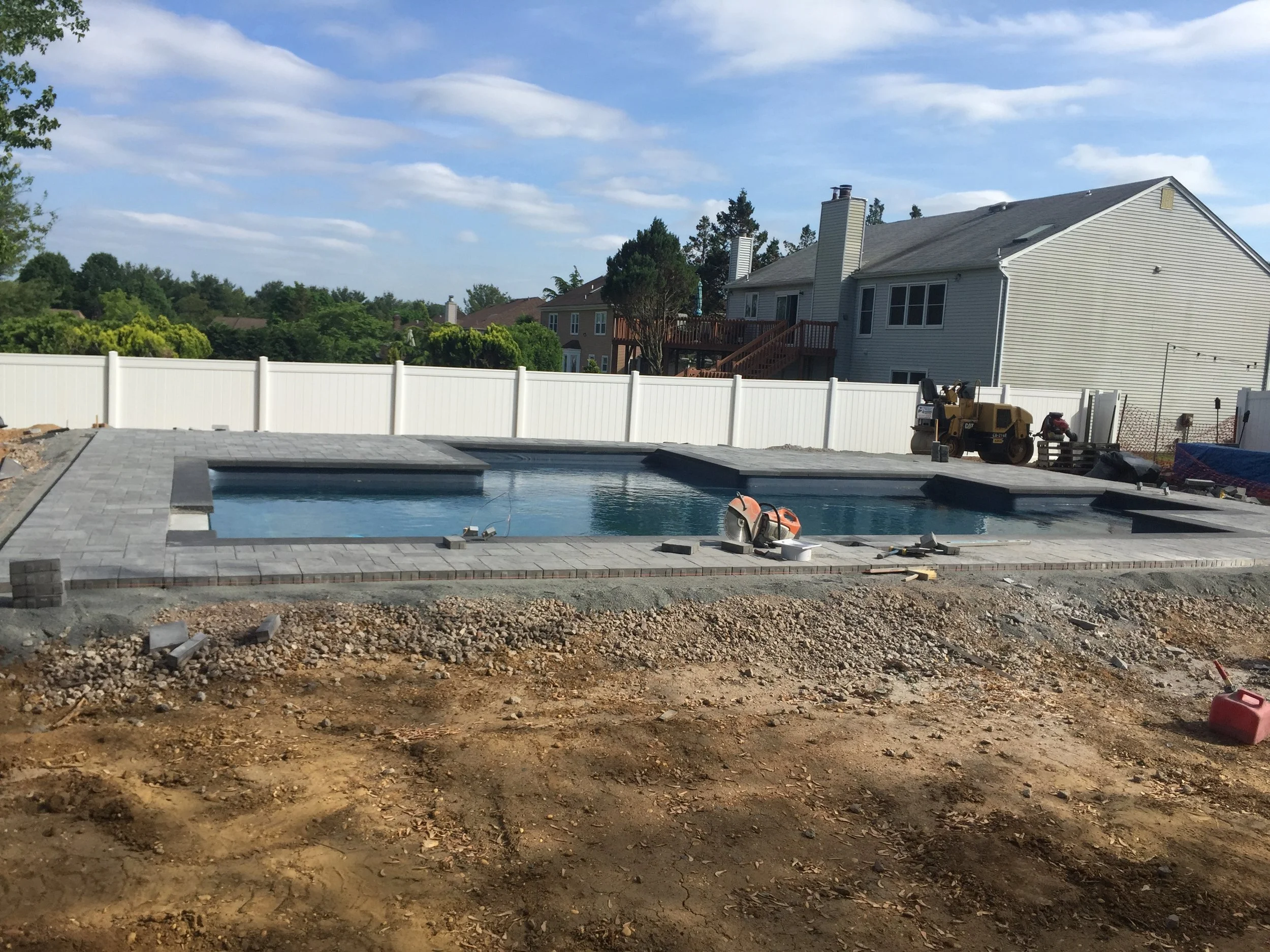 A swimming pool under construction in a backyard, with a white fence, a house with a deck, and trees in the background. Construction tools and equipment are visible around the pool area.