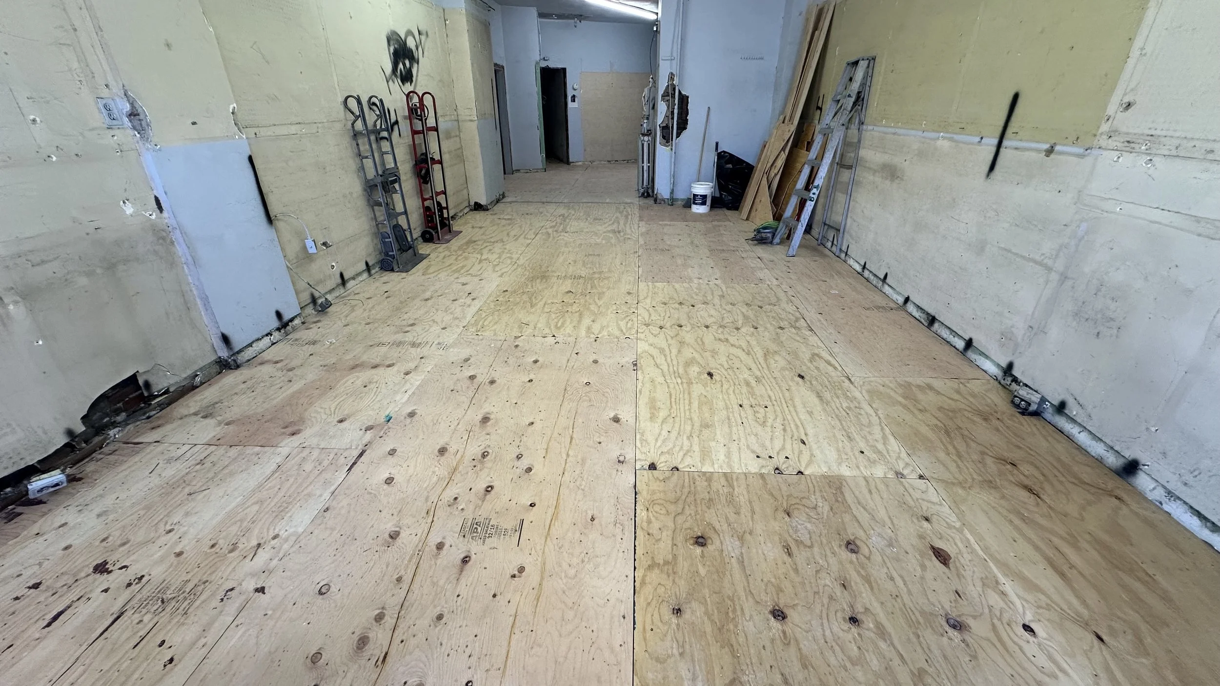 Room under construction with plywood flooring, ladders, and construction tools along the walls.
