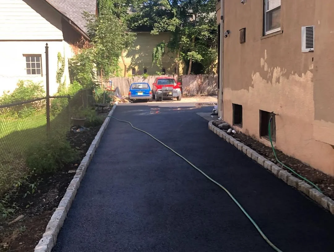 A newly paved asphalt driveway with a garden hose lying on it, bordered by brick edging, leading to a backyard with parked cars and a wooden fence.