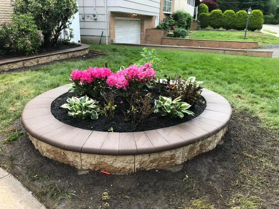 A circular flower bed with a brick border, filled with pink and white flowers, in a suburban front yard.