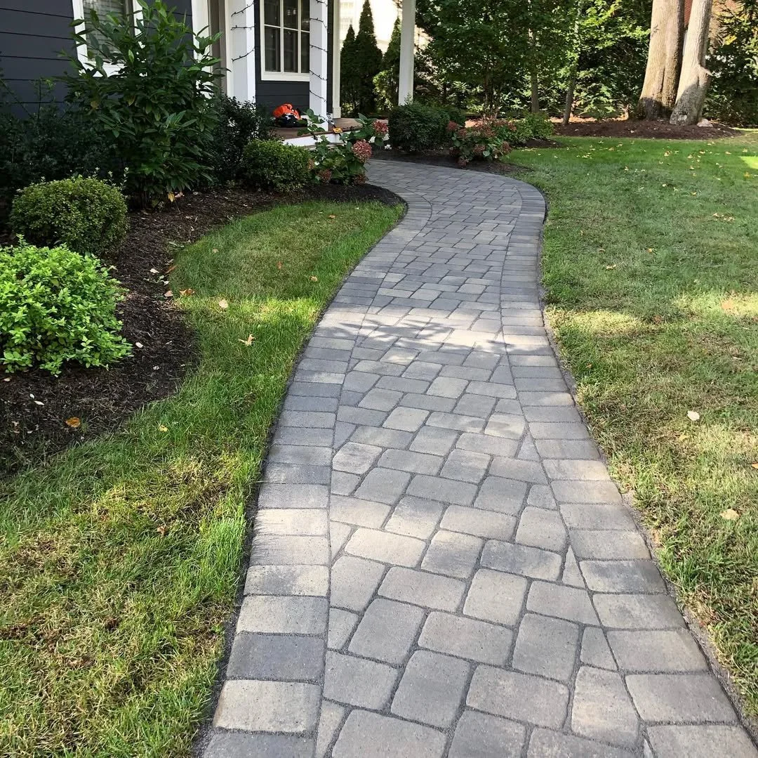 A curved gray brick pathway leading to a house with white siding and a porch, surrounded by green grass, bushes, and trees.