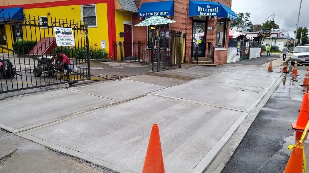 Concrete sidewalk and driveway in front of a restaurant, with orange traffic cones and construction barriers.
