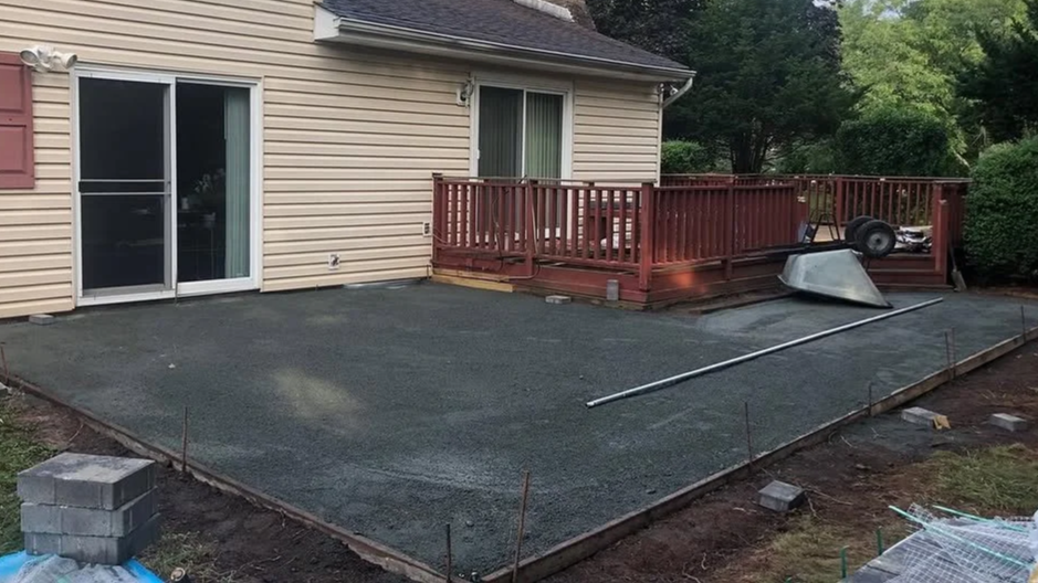 A backyard patio under construction with a newly laid gravel surface, a wooden deck with a railing, and construction tools including a wheelbarrow, a metal pipe, and concrete blocks. The area is bordered by wooden framing and adjacent to a house with sliding glass doors.