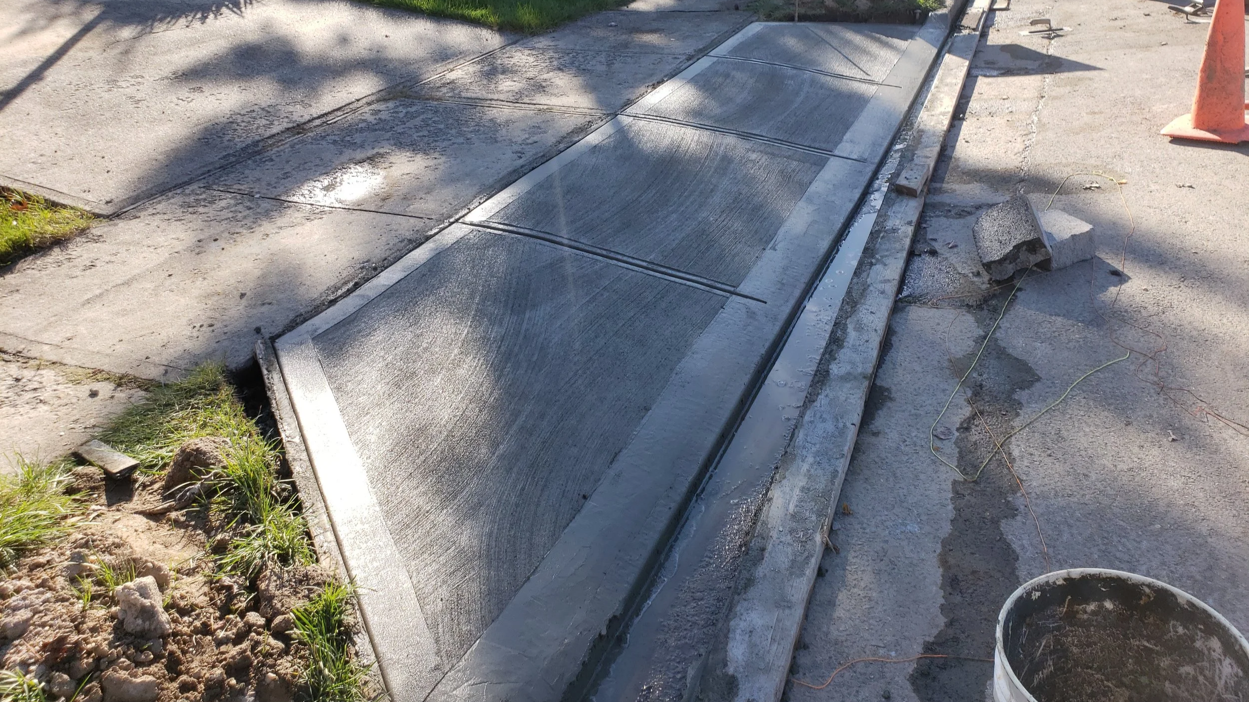 A sidewalk curb with new concrete being poured and finished, with some construction debris and a traffic cone nearby.