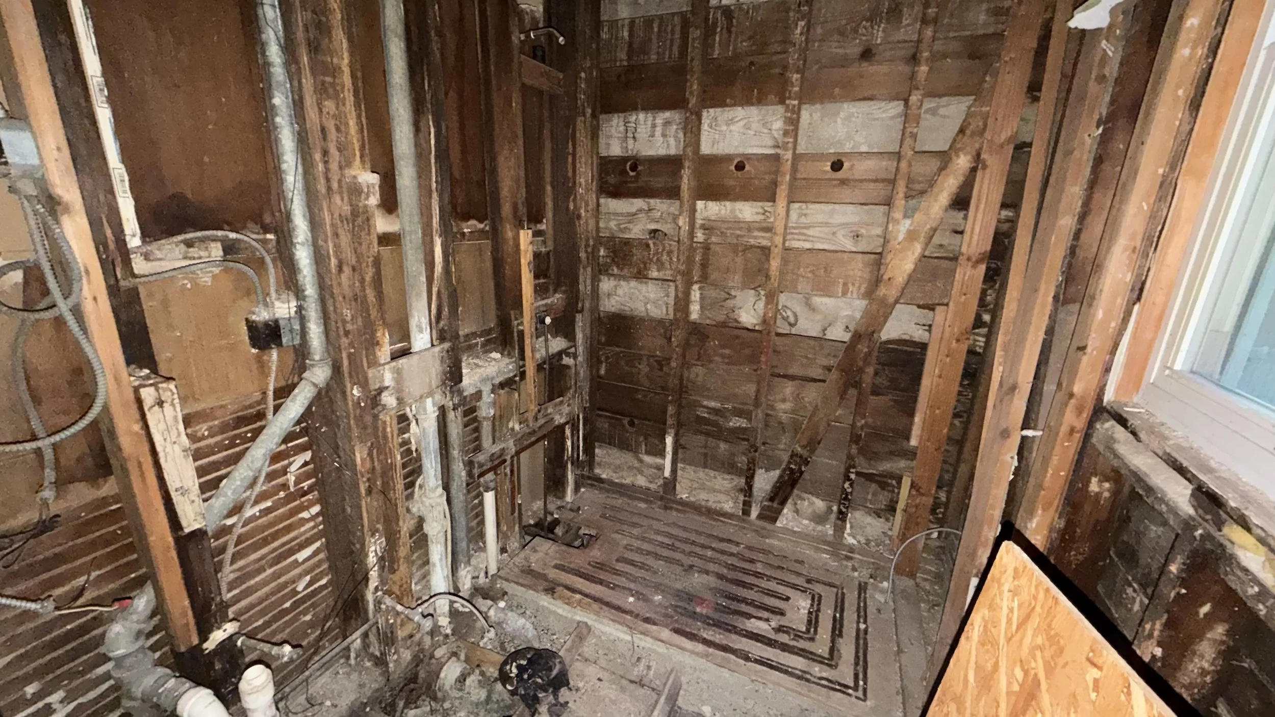 Interior of a room under demolition with exposed wooden framing, pipes, and electrical wiring visible.
