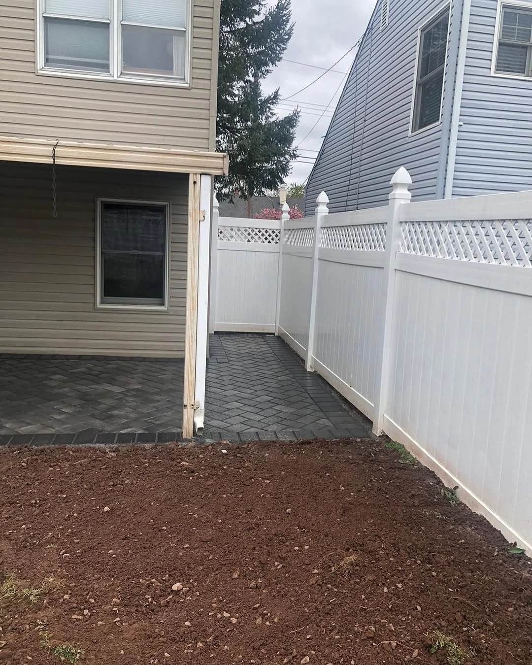 Backyard with new white vinyl fence, paved pathway, and soil area with neighboring houses.