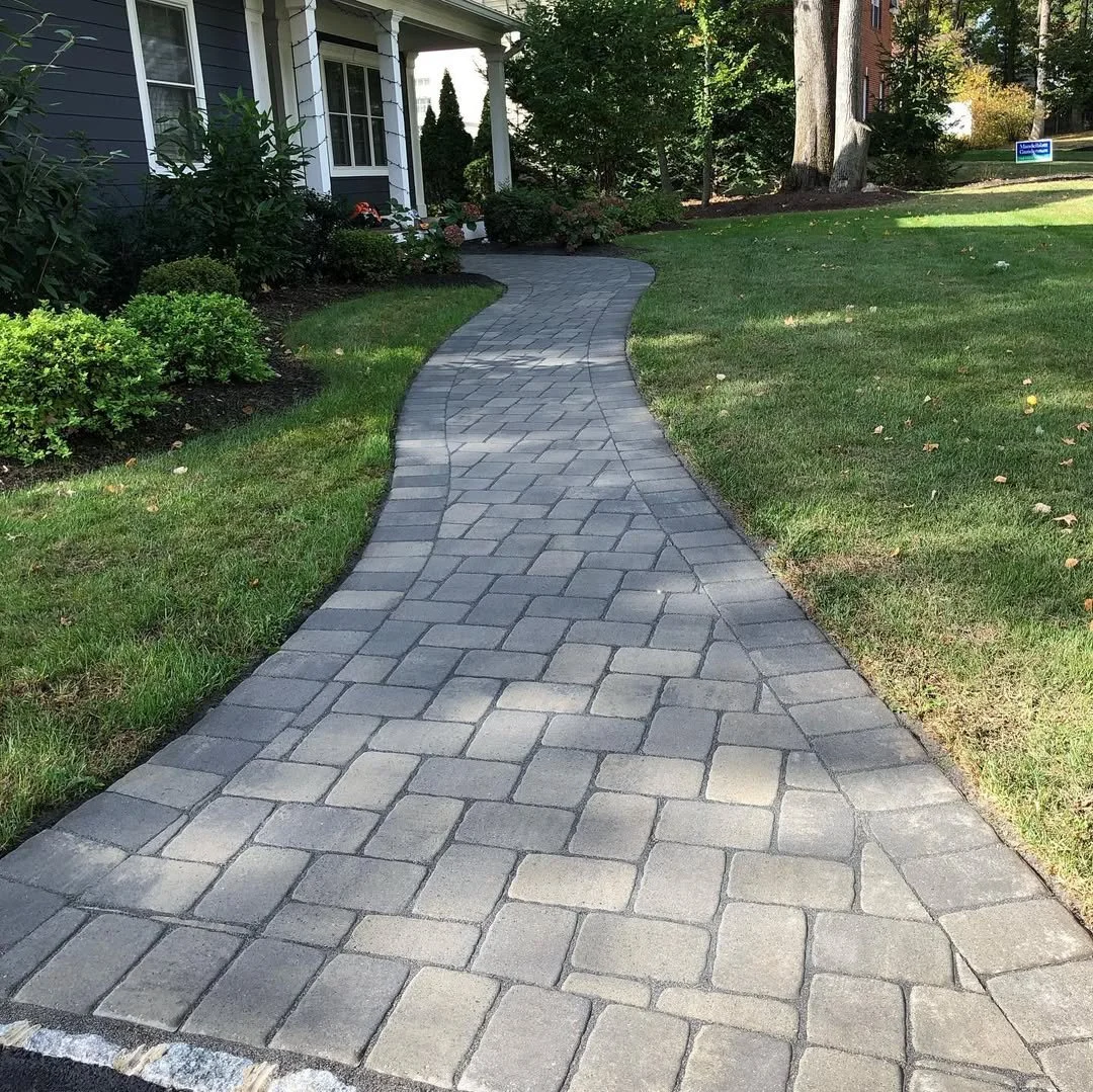A curved stone walkway leads to a house with a porch, surrounded by green lawn and bushes, with trees in the background.