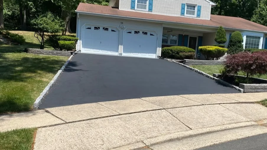 A freshly paved black asphalt driveway leading up to a white double garage with decorative windows, bordered by garden beds with shrubs and bushes, in front of a light-colored house with blue shutters.