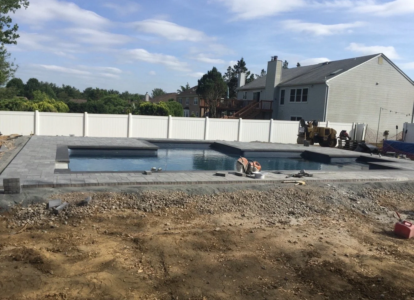 A backyard swimming pool under construction with a fence around it, with construction equipment and building materials nearby, and houses in the background.