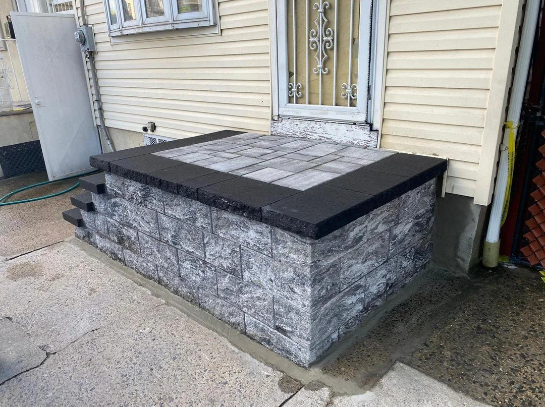 Concrete porch or step with a stone wall base, topped with a surface made of paving stones, and an inset of dark bricks around the top edge, adjacent to a house with beige siding and a door with a white security screen.