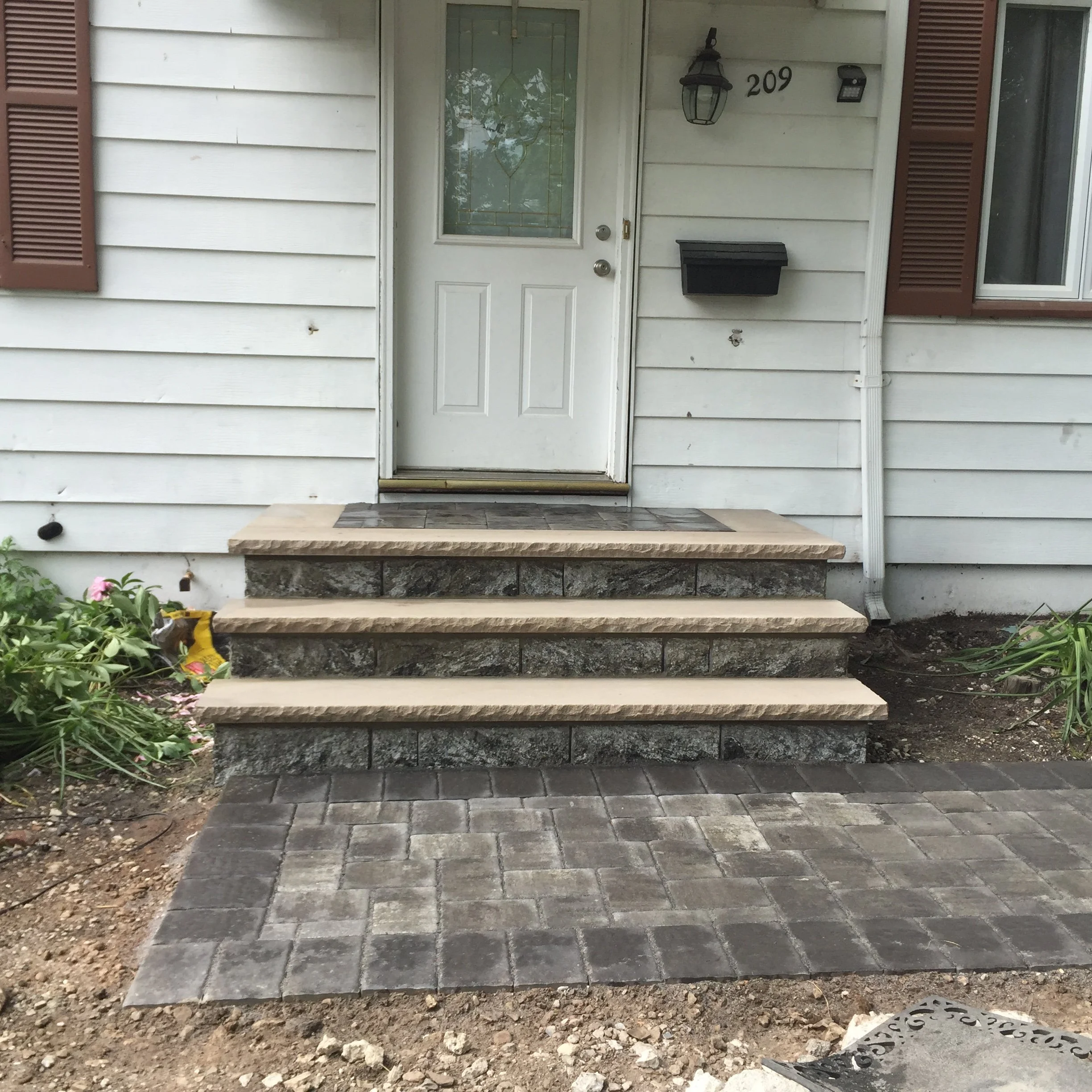 Front steps leading to a house with a white door, black mailbox, and house number 209. There are three steps made of stone with a beige edging, a small porch area paved with gray bricks, and some plants on either side of the steps.