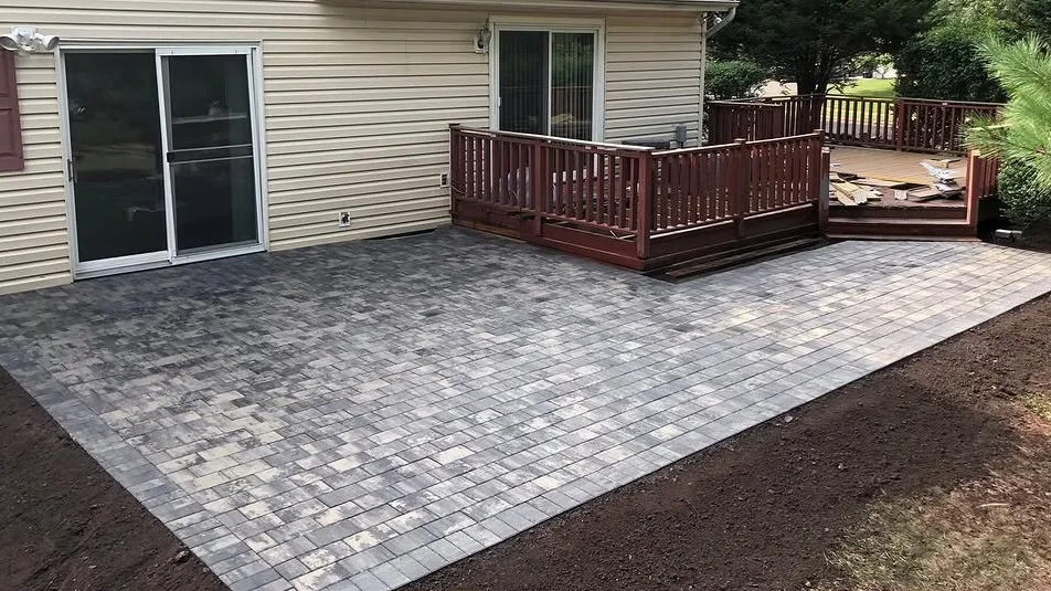 Newly installed paver patio with a small wooden deck attached to a house with sliding glass doors and a window, surrounded by bare soil and green trees in the background.