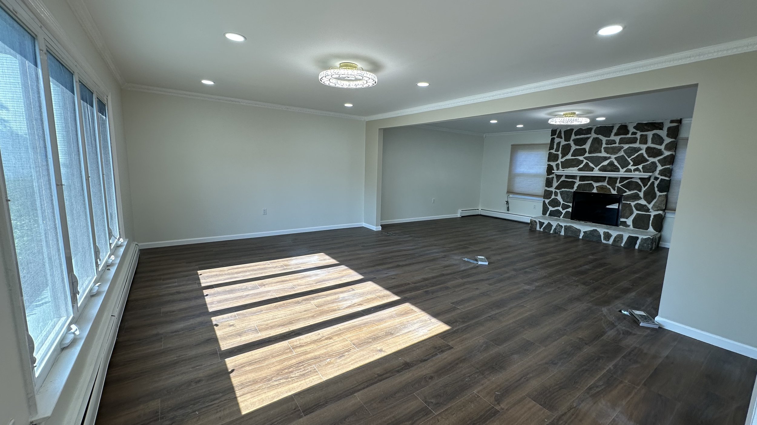 Empty living room with wood flooring, large windows letting in sunlight, a stone fireplace, and neutral wall colors.