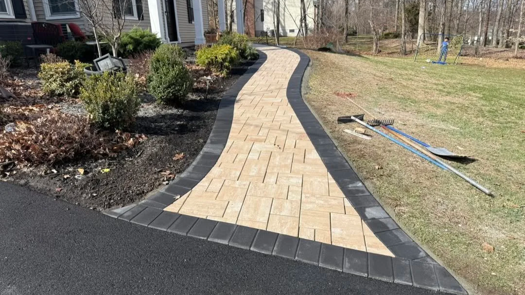 Curved walkway made of beige pavers bordered by black bricks, leading past a garden bed with bushes and an outdoor house in the background, with gardening tools on the grass to the right.